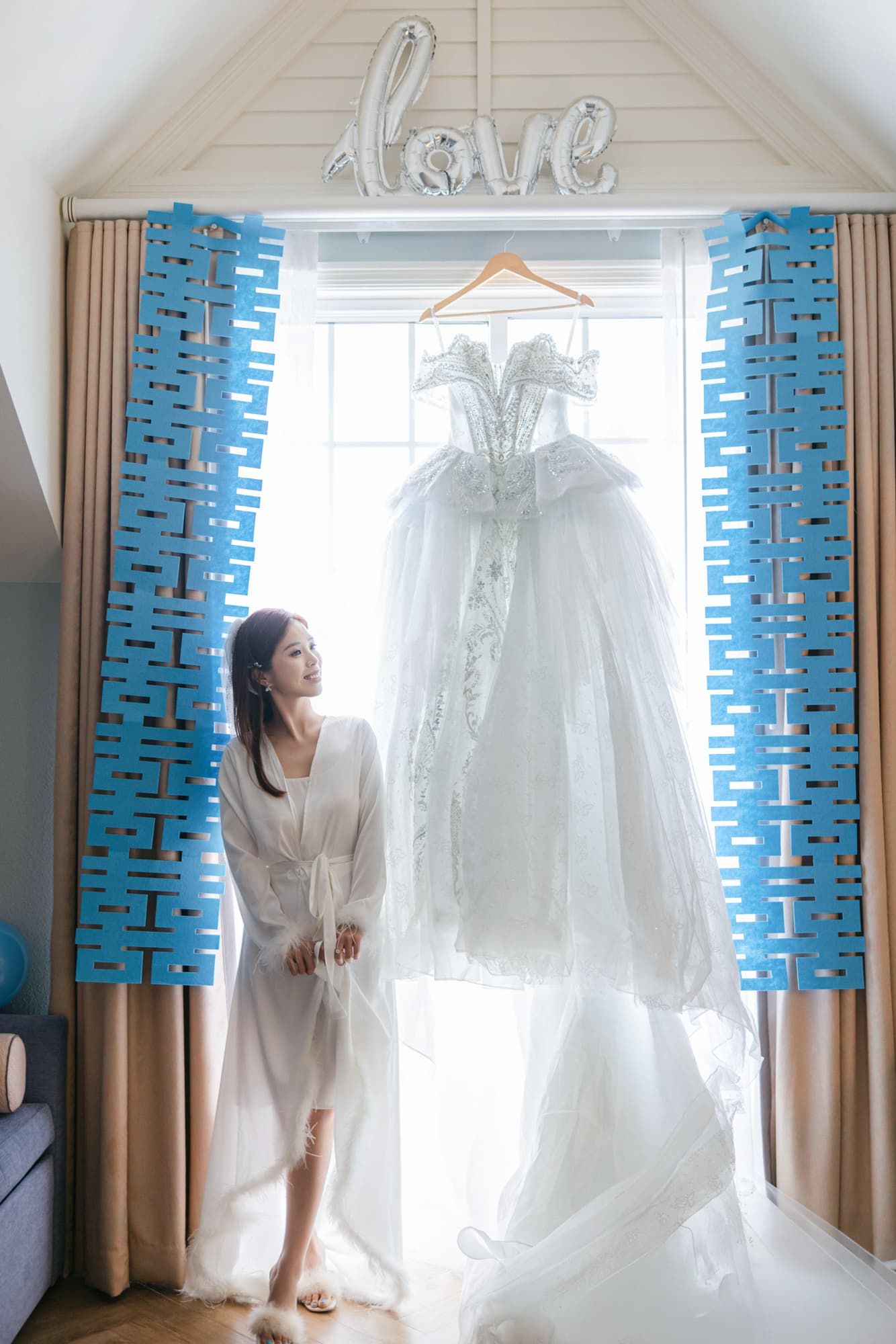 Bride in white robe admiring her lace wedding dress hanging by sunlit window with love balloon.