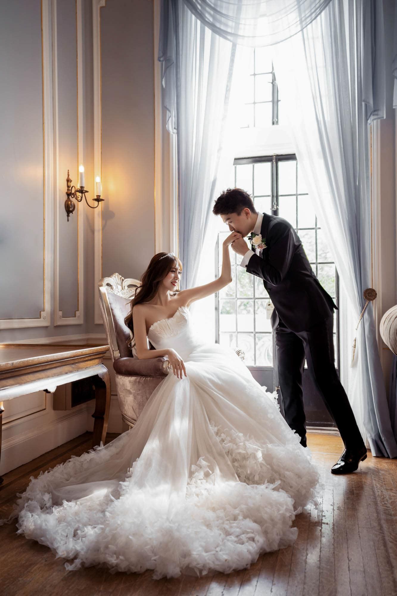 Bride in a voluminous gown sits in a chair as groom kisses her hand by a tall window.