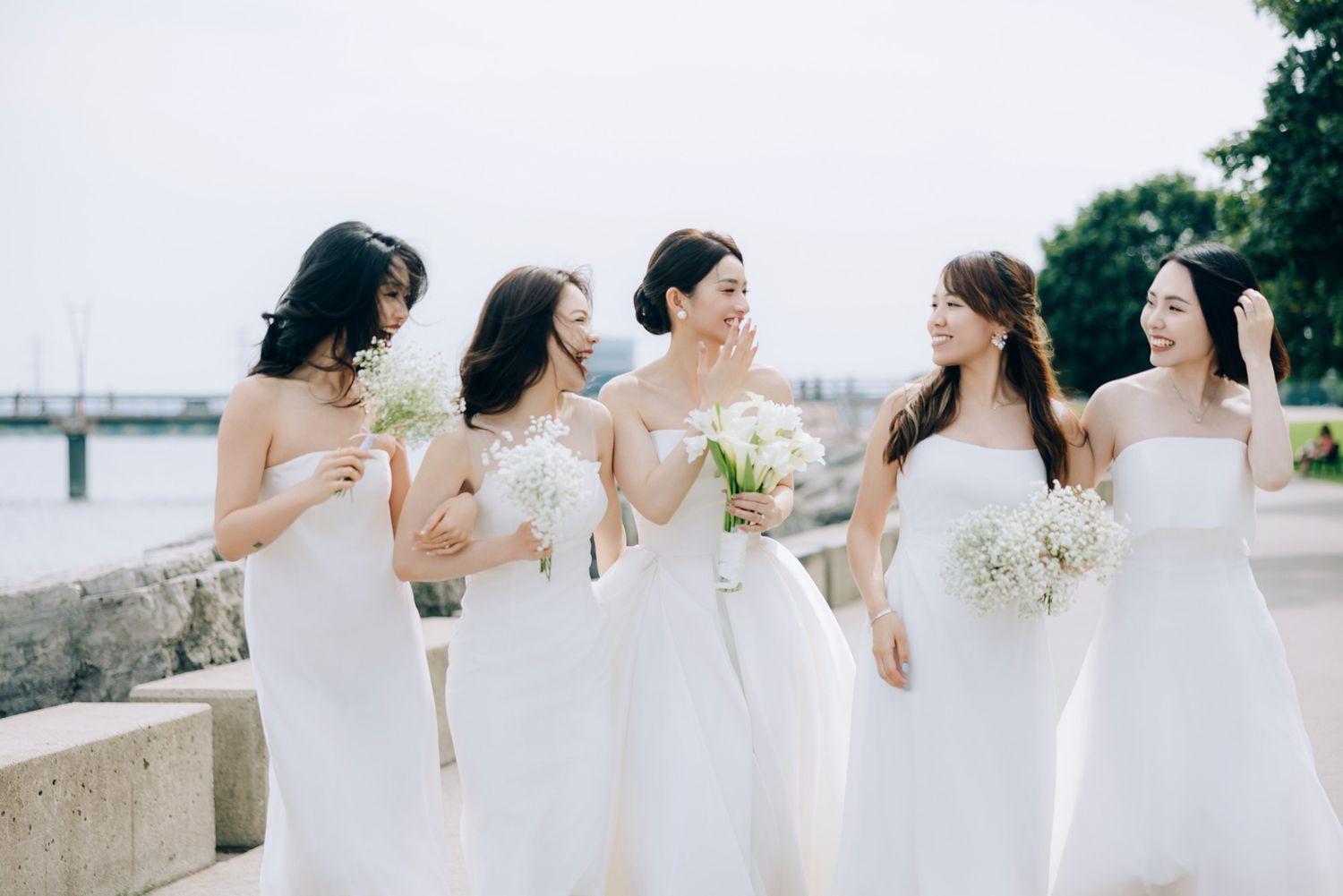 Five bridesmaids in matching white dresses walk together holding white floral bouquets.