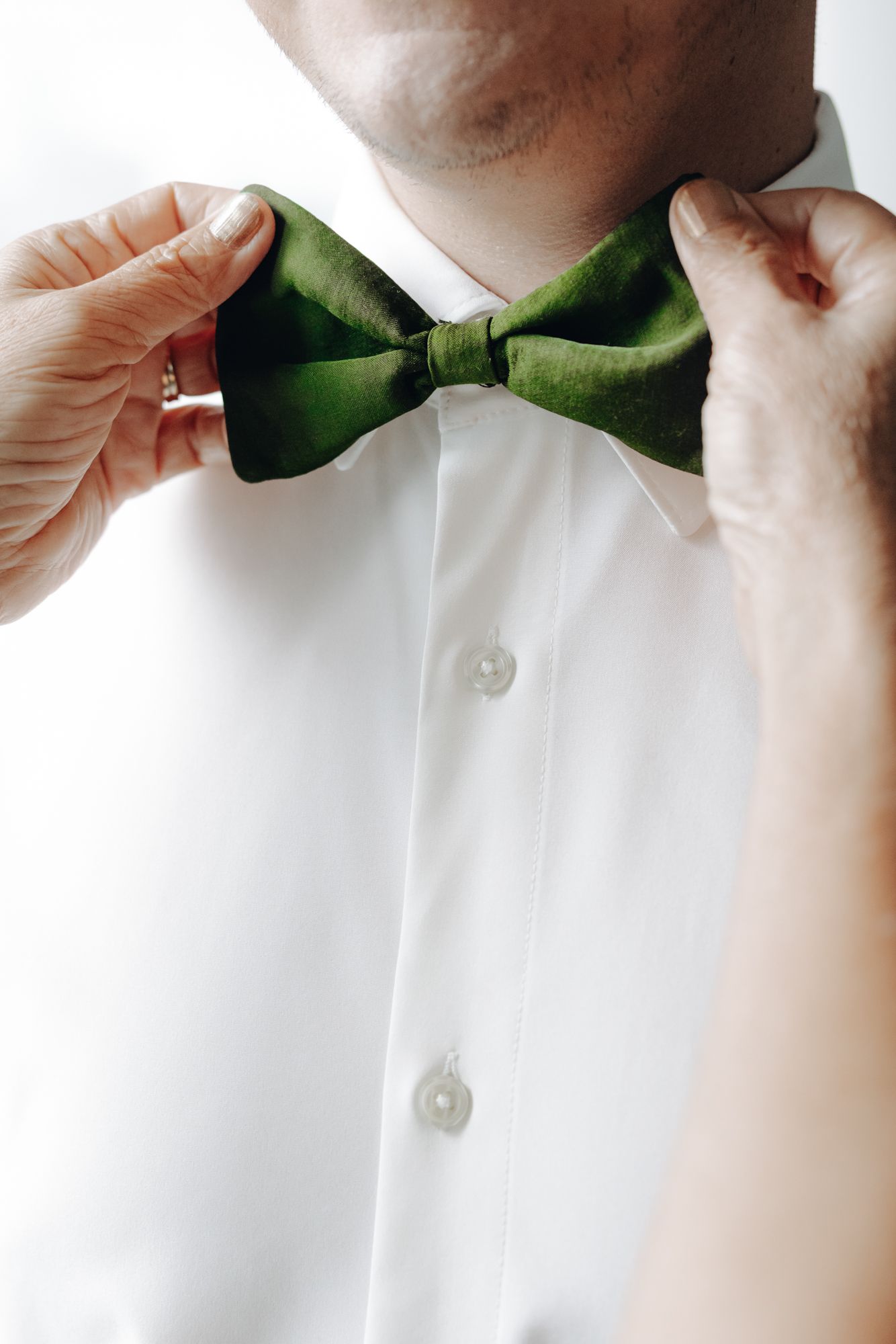 Two hands adjusting a green silk bow tie on a man wearing a crisp white dress shirt with pearl buttons.