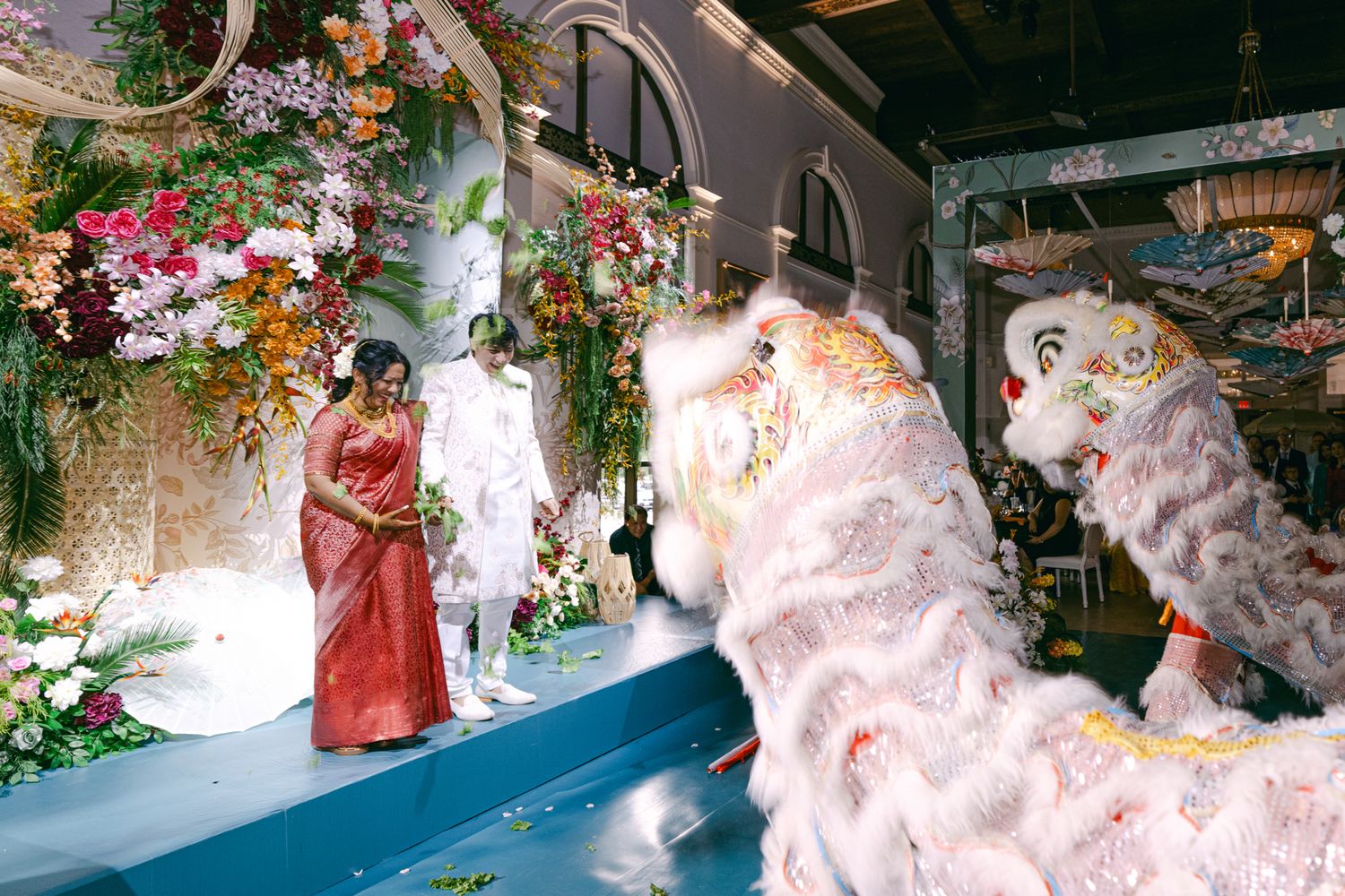 Bride in red dress and groom in white stand on a stage near floral arrangements as two white lion dancers perform at a wedding.