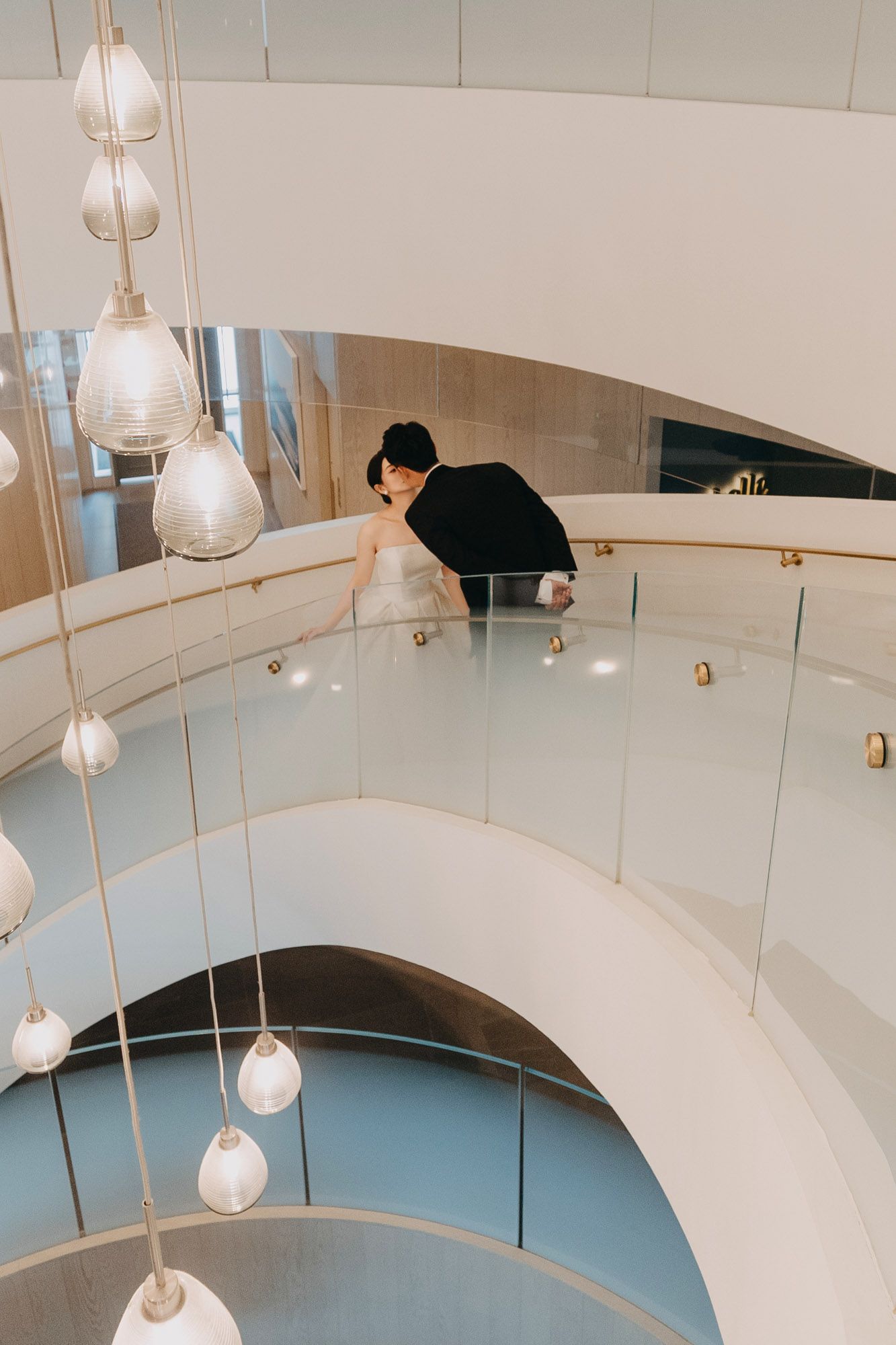 Bride and groom share a romantic moment on a curved glass balcony of a modern white spiral staircase.