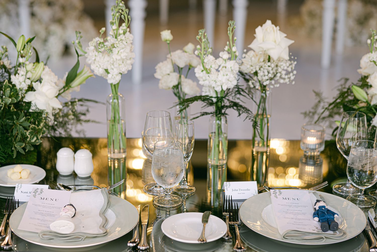 An elegant wedding reception table with white floral centerpieces, glassware, place settings, and menu cards on a mirrored surface.