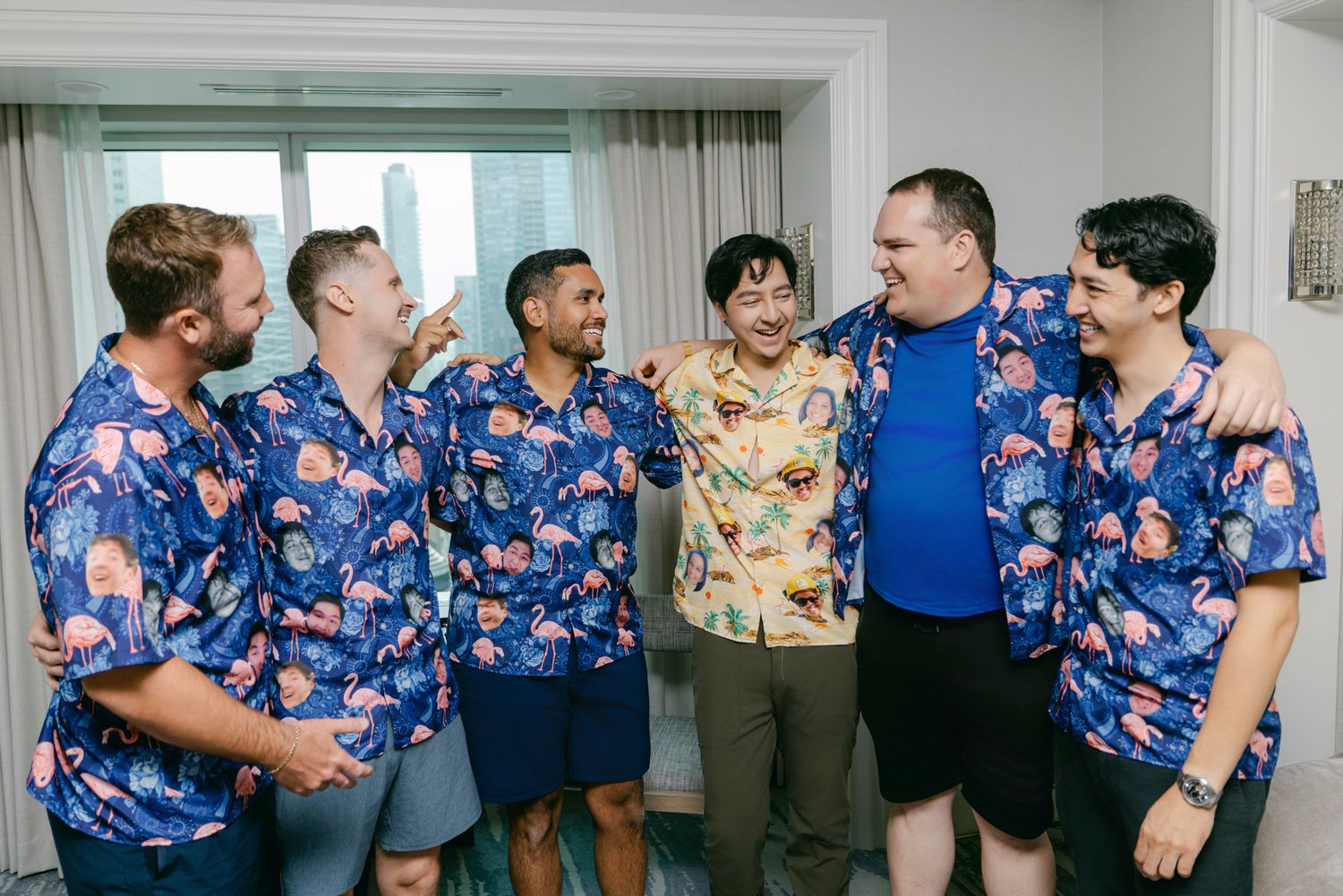 Groomsmen in matching floral Hawaiian shirts laugh and pose together with their arms around each other in a bright hotel room.