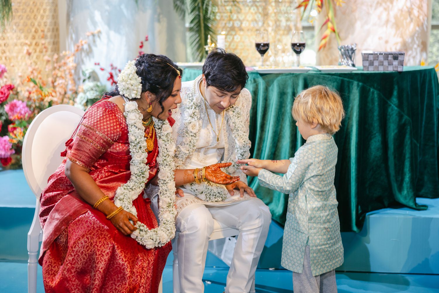 Bride in red silk sari and groom in white garlands sit at a Hindu wedding ceremony as a young child hands them a small item.