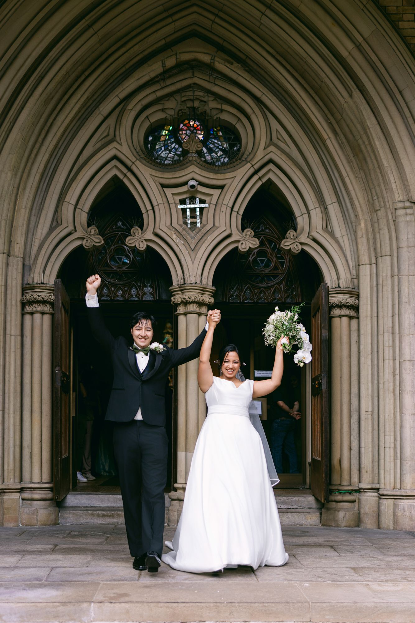Joyful newlyweds raise their arms in celebration as they exit a Gothic stone church, the bride in a white gown holding a bouquet.