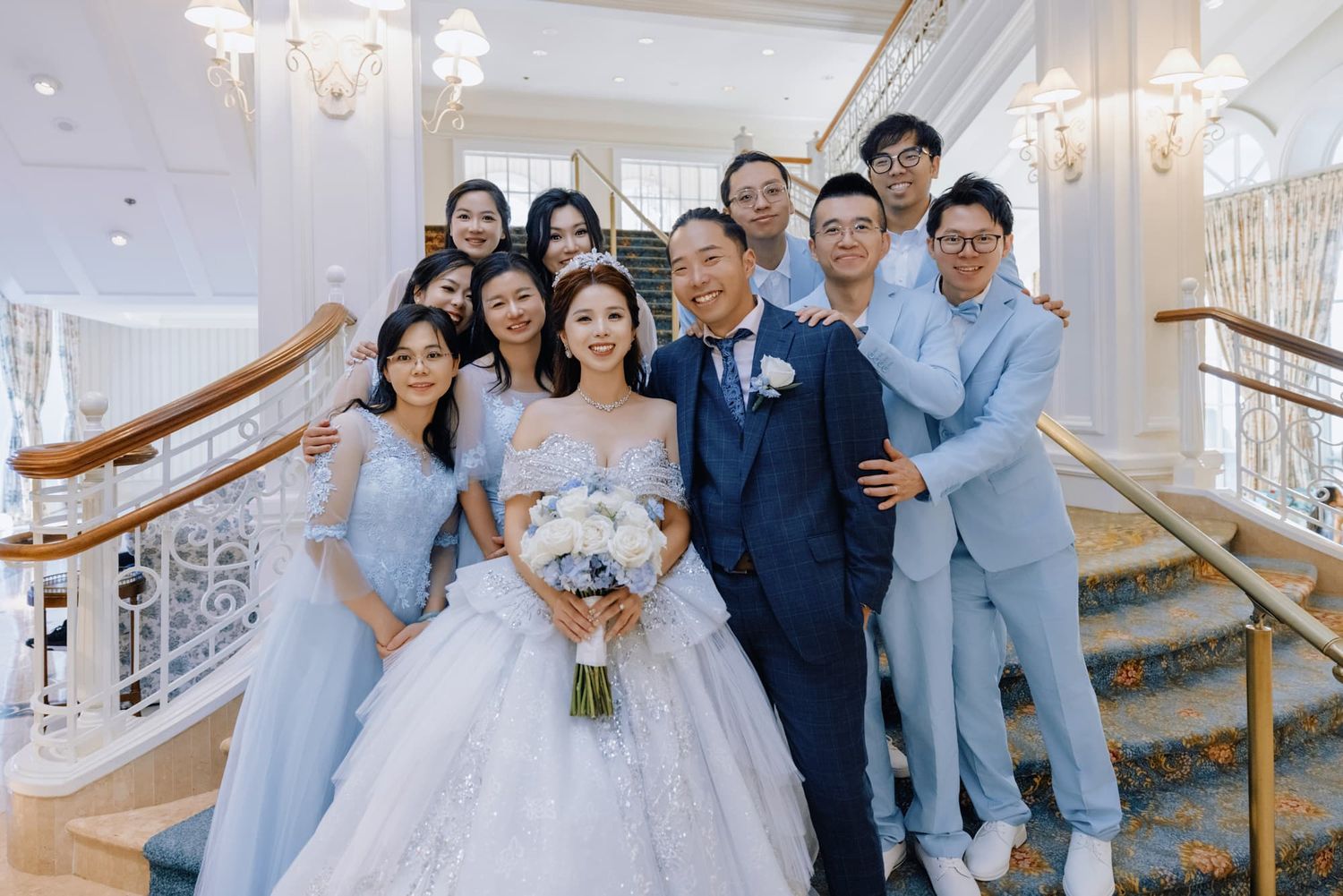 Bride in white ball gown holds bouquet with wedding party in blue attire on elegant staircase.