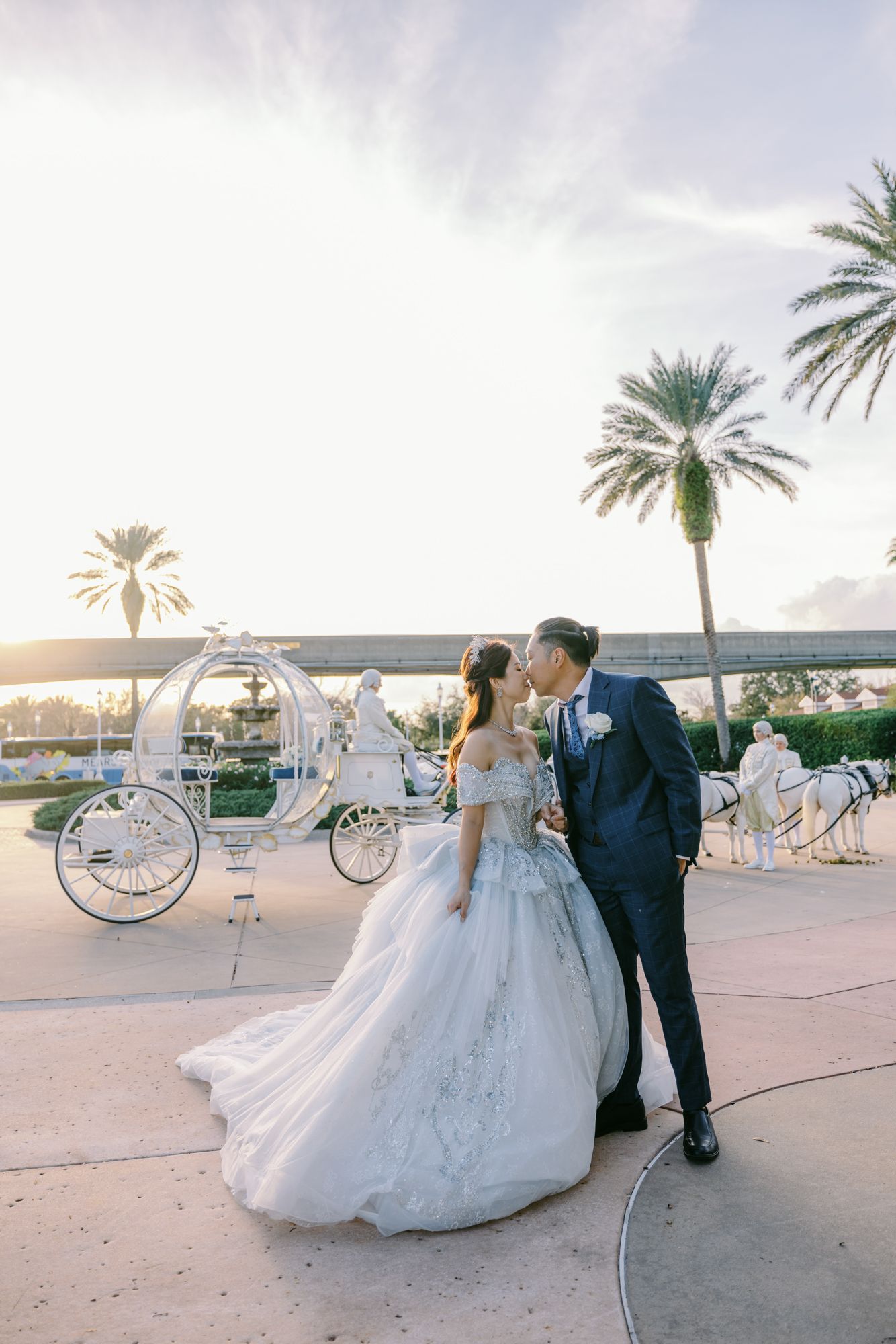 Bride and groom kissing near Cinderella carriage with palm trees at sunset in Disney.