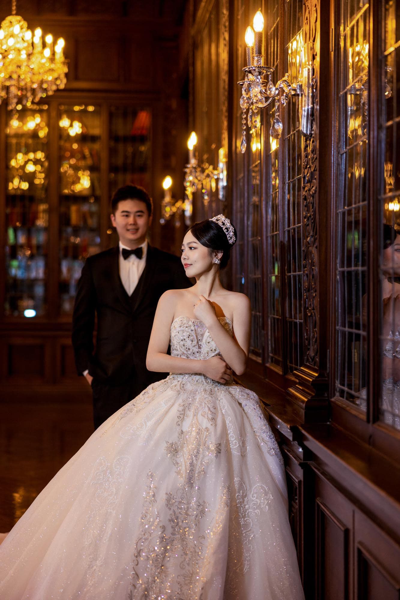 Bride in embellished ballgown with tiara and groom in tuxedo posing in a warmly lit ornate library.