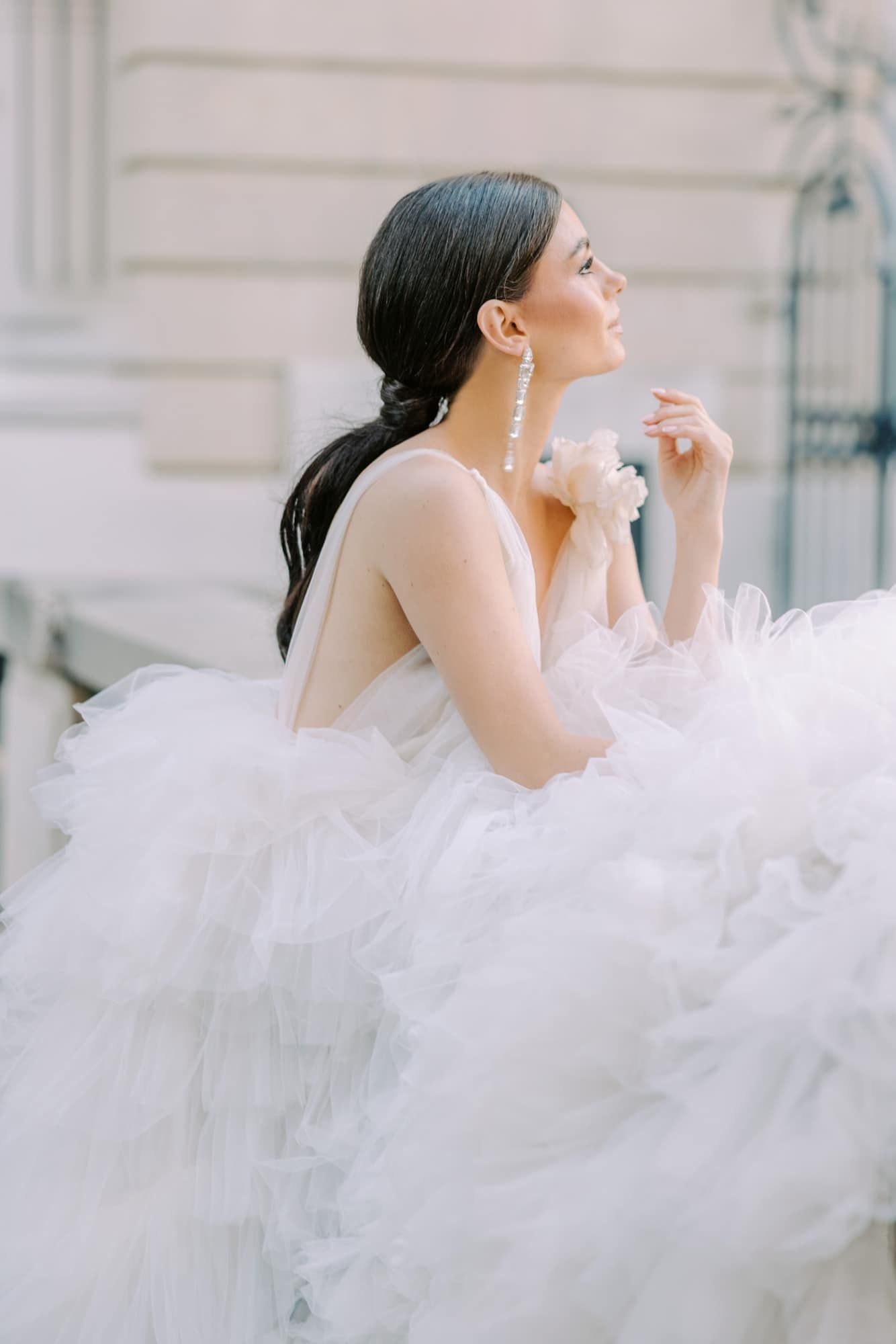 Elegant bride in a voluminous white tulle wedding gown with pearl drop earrings, sitting gracefully on outdoor steps.