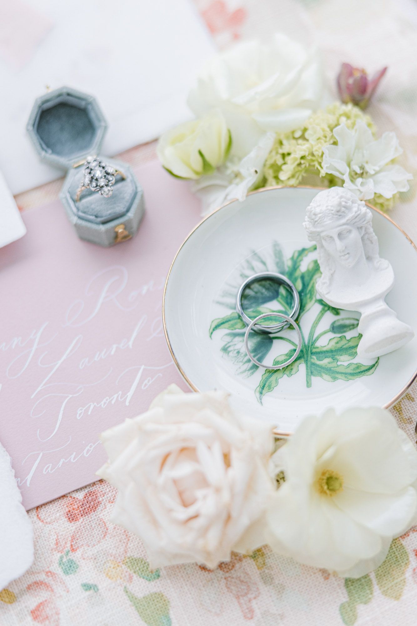 Elegant wedding flat lay featuring diamond ring, gold bands, white roses, and calligraphy invitation.
