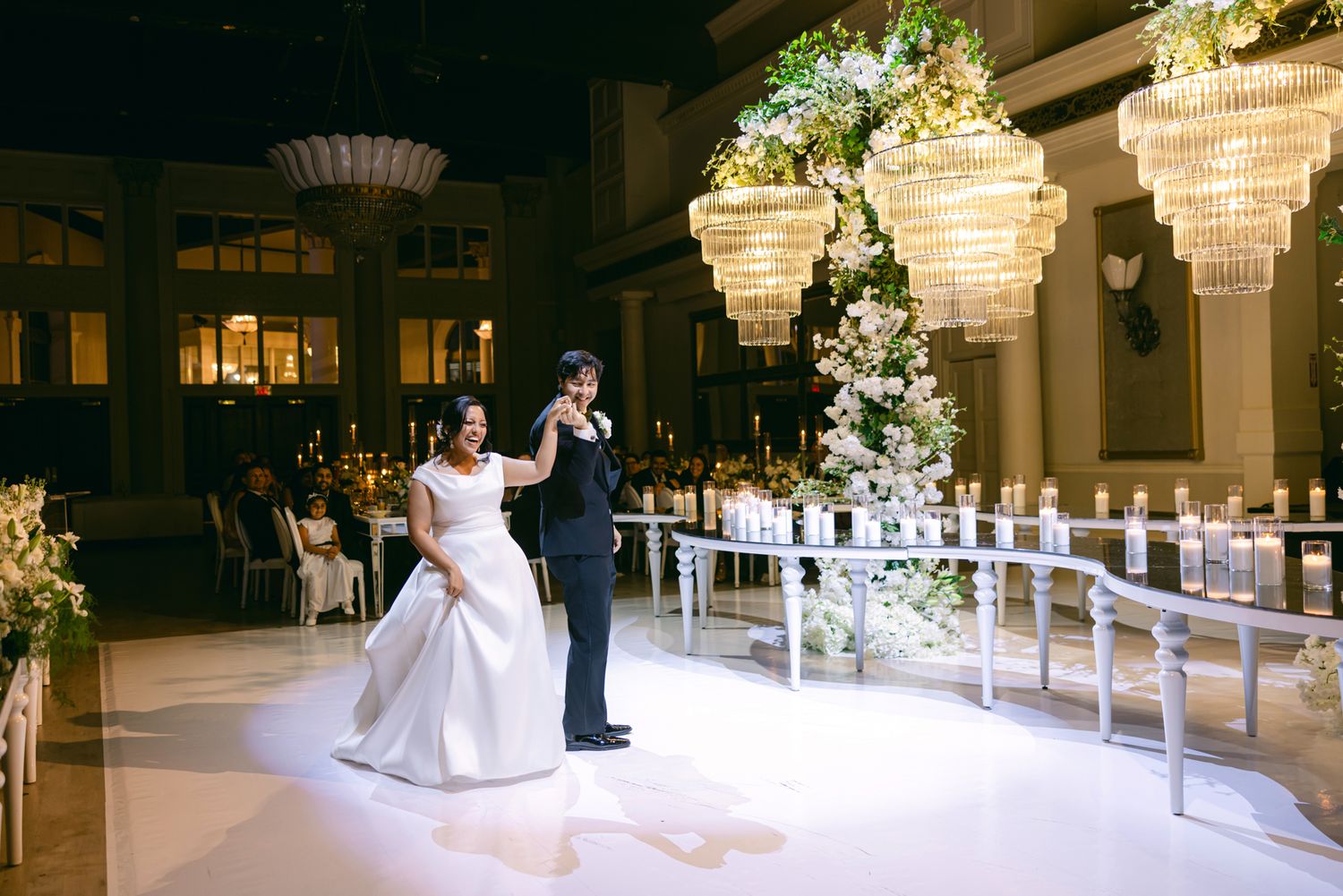 Bride in a white ball gown and groom in a black tuxedo share a first dance in a ballroom with crystal chandeliers.
