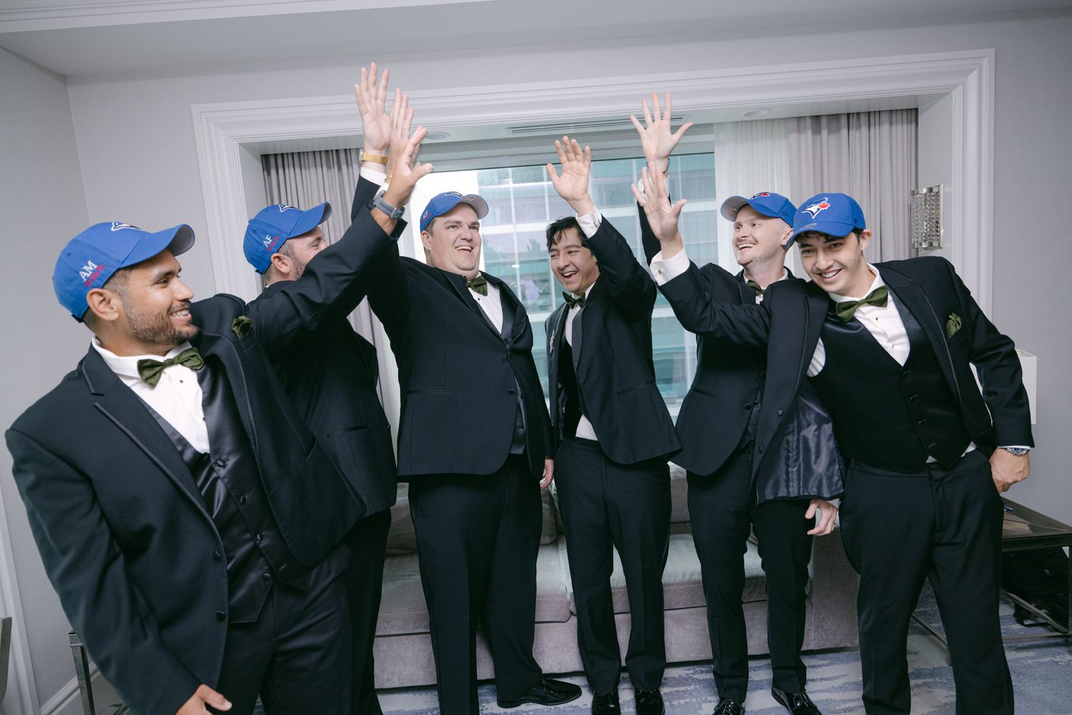 Groom and groomsmen in black tuxedos and blue baseball caps raise their hands and cheer together in a hotel room.