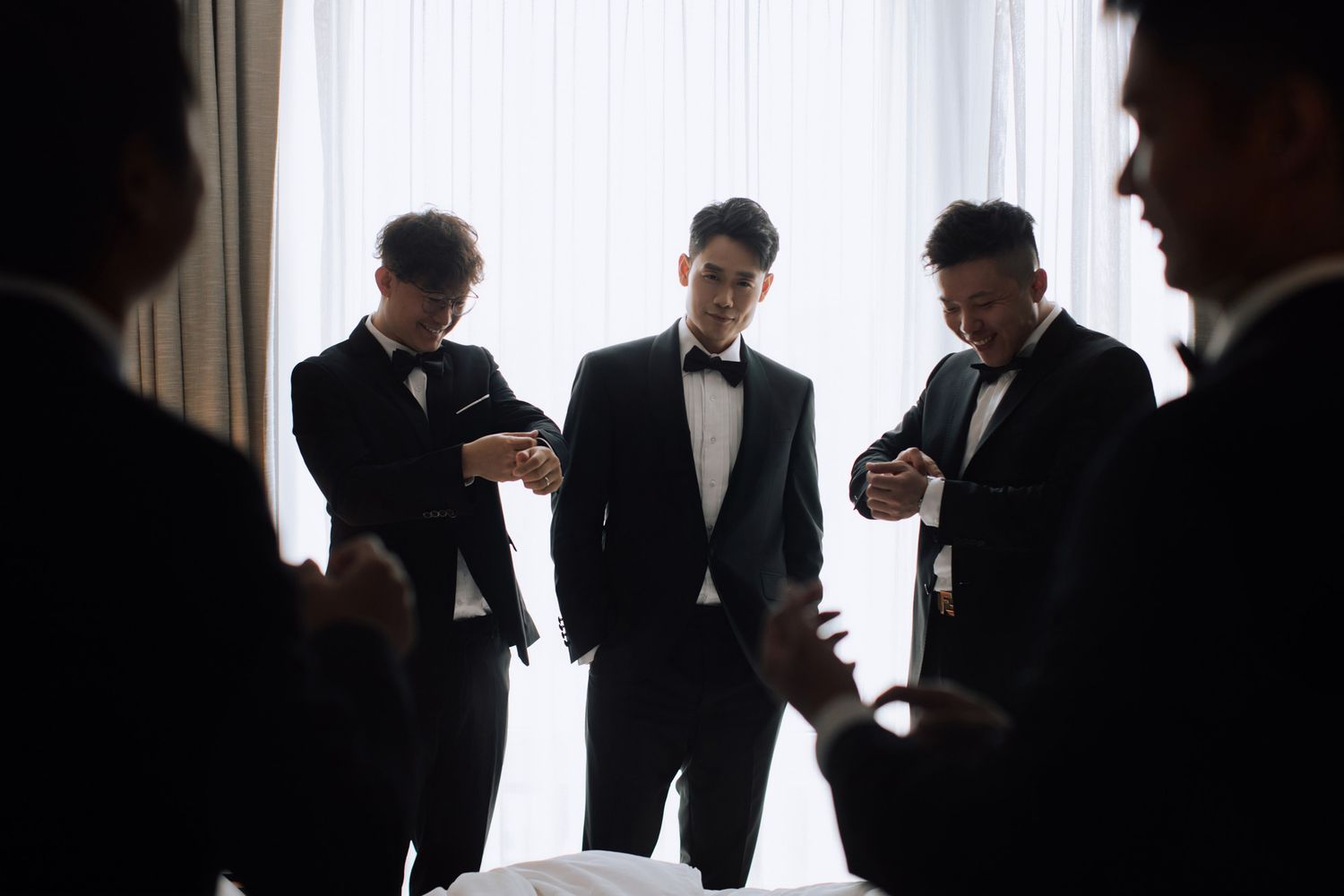 Groom and groomsmen in black tuxedos checking cufflinks and details before wedding ceremony.