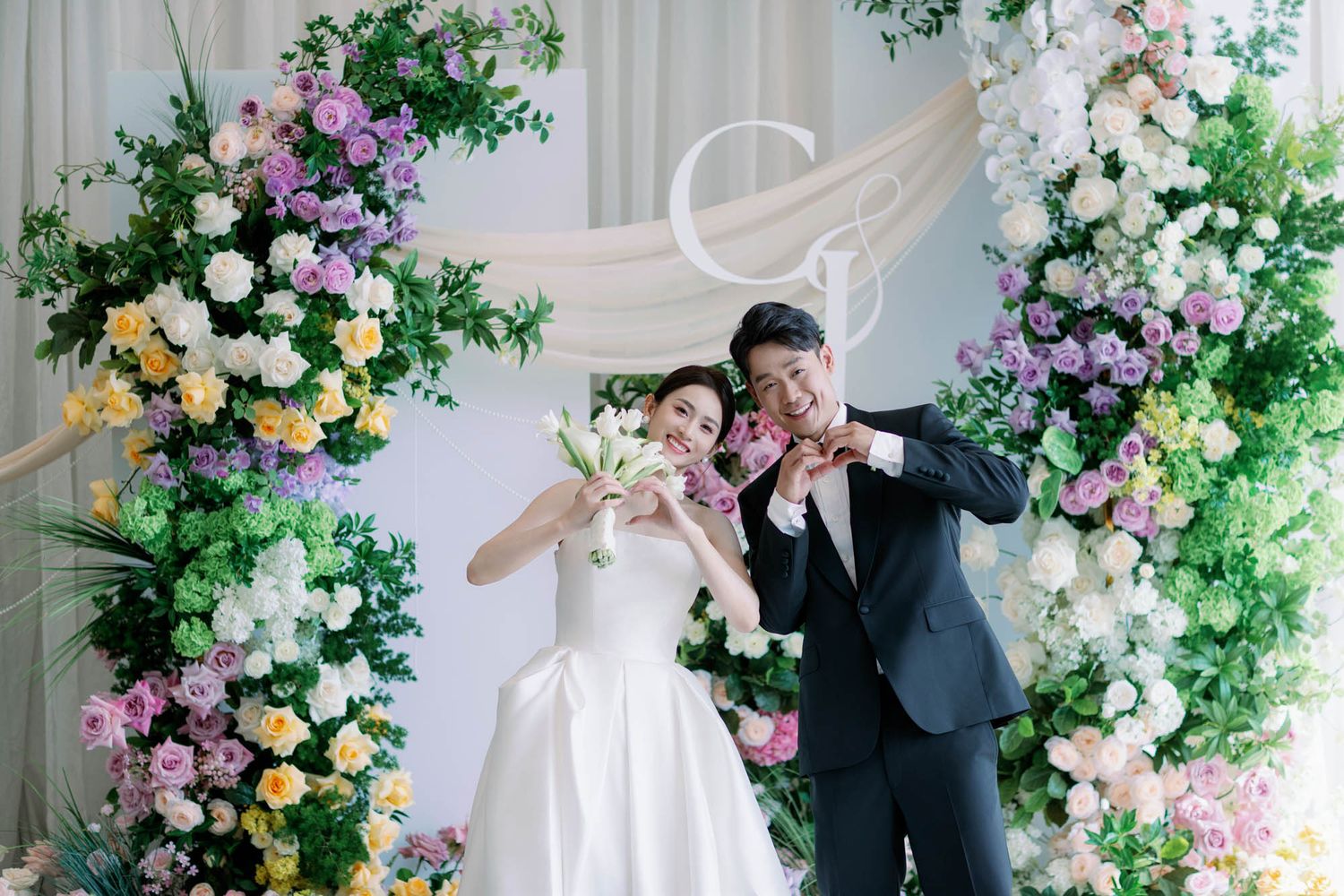 Happy couple making heart gestures in front of a beautiful floral wedding arch with pastel flowers.