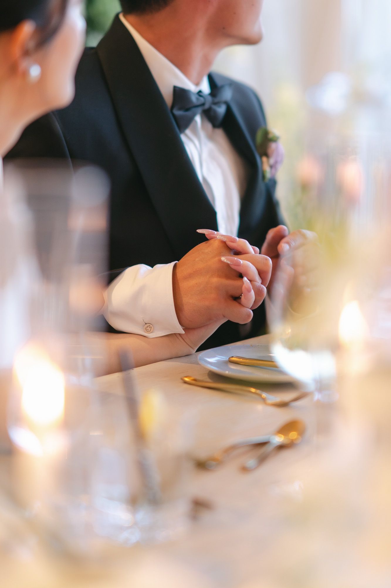 Close-up of bride and groom holding hands at wedding reception table with candles and gold cutlery.