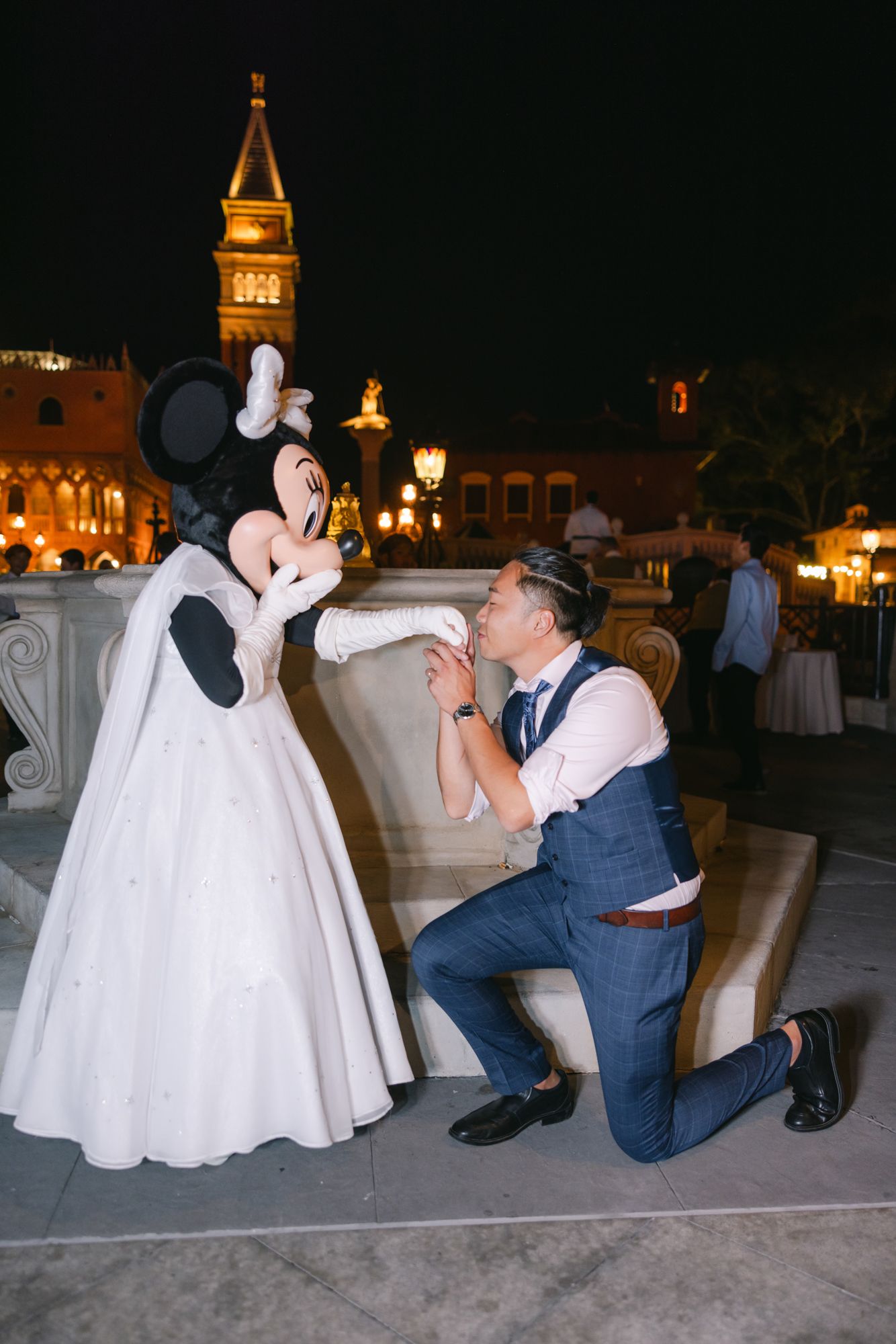 Groom kneels playfully kissing Minnie Mouse character hand at Disney wedding reception at night.