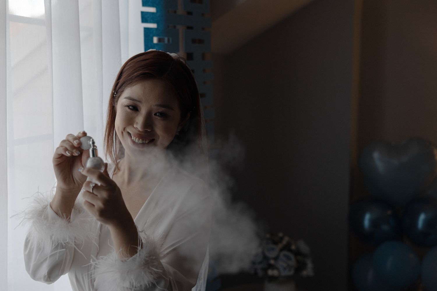 Smiling Asian woman in white robe holding perfume bottle with mist cloud indoors.
