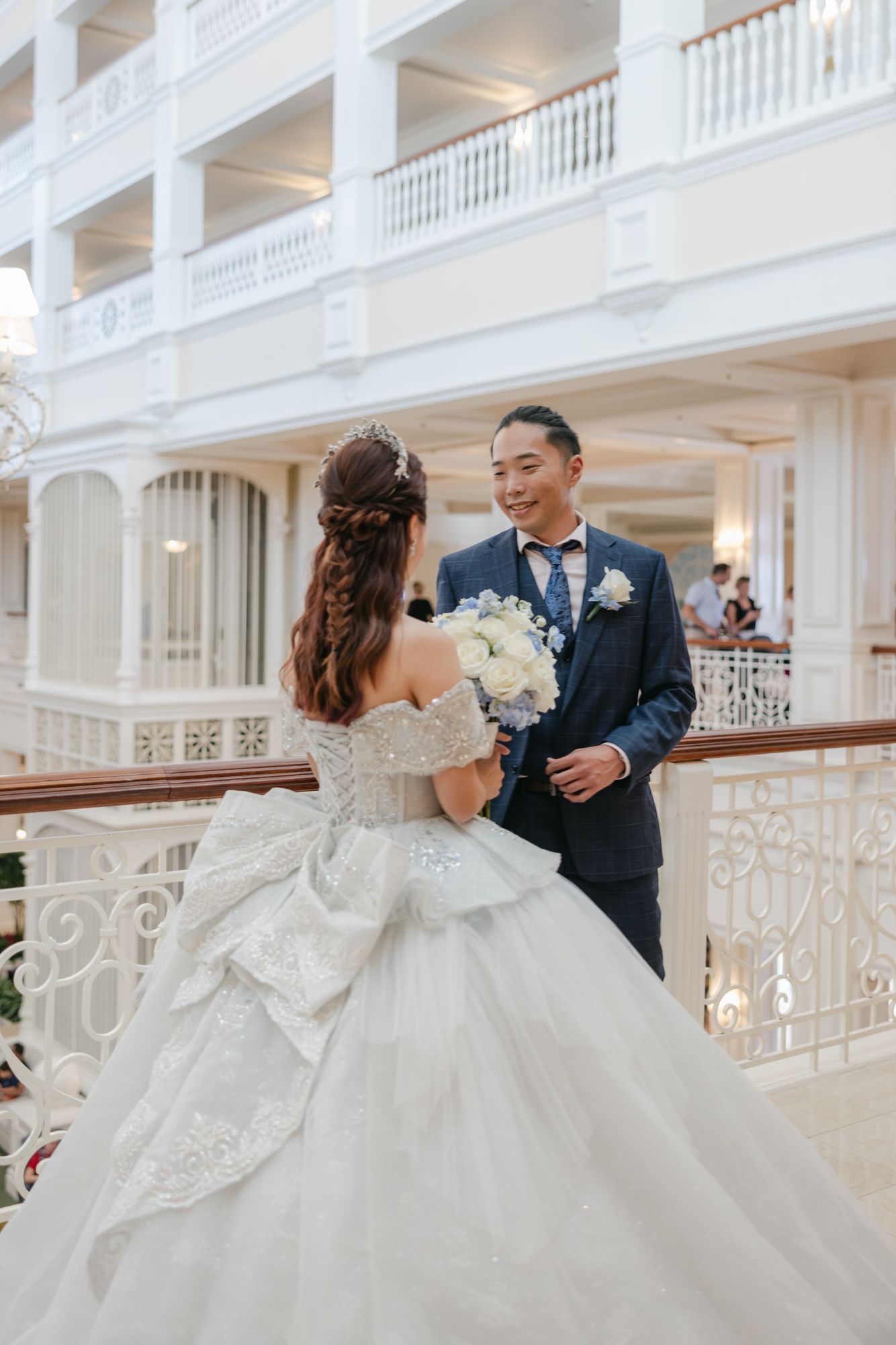 Bride in elegant lace gown holds bouquet facing groom in navy suit on grand white hotel balcony.