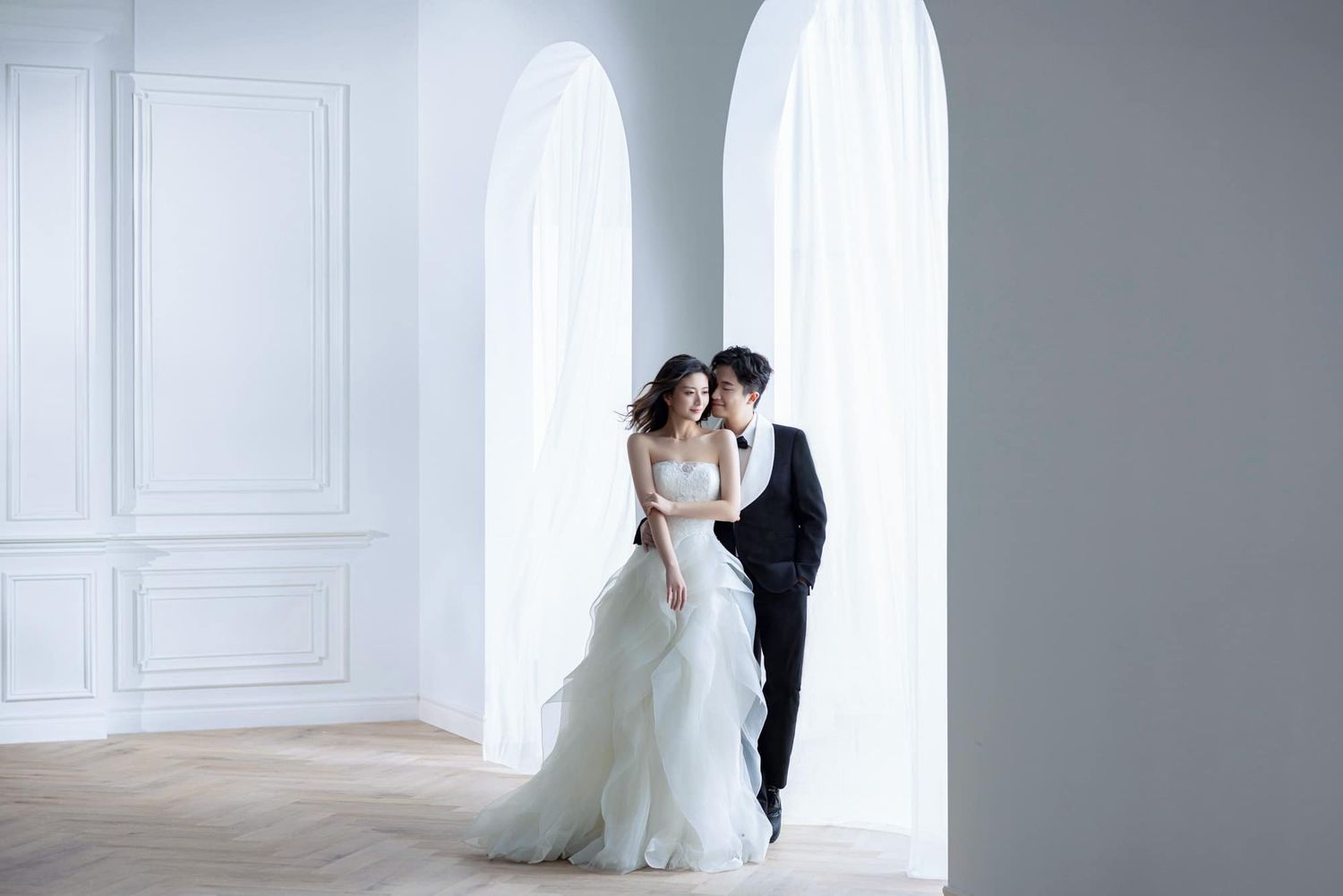 Elegant bride in white gown and groom in black tuxedo posing together in a bright, minimalist studio for wedding photos.