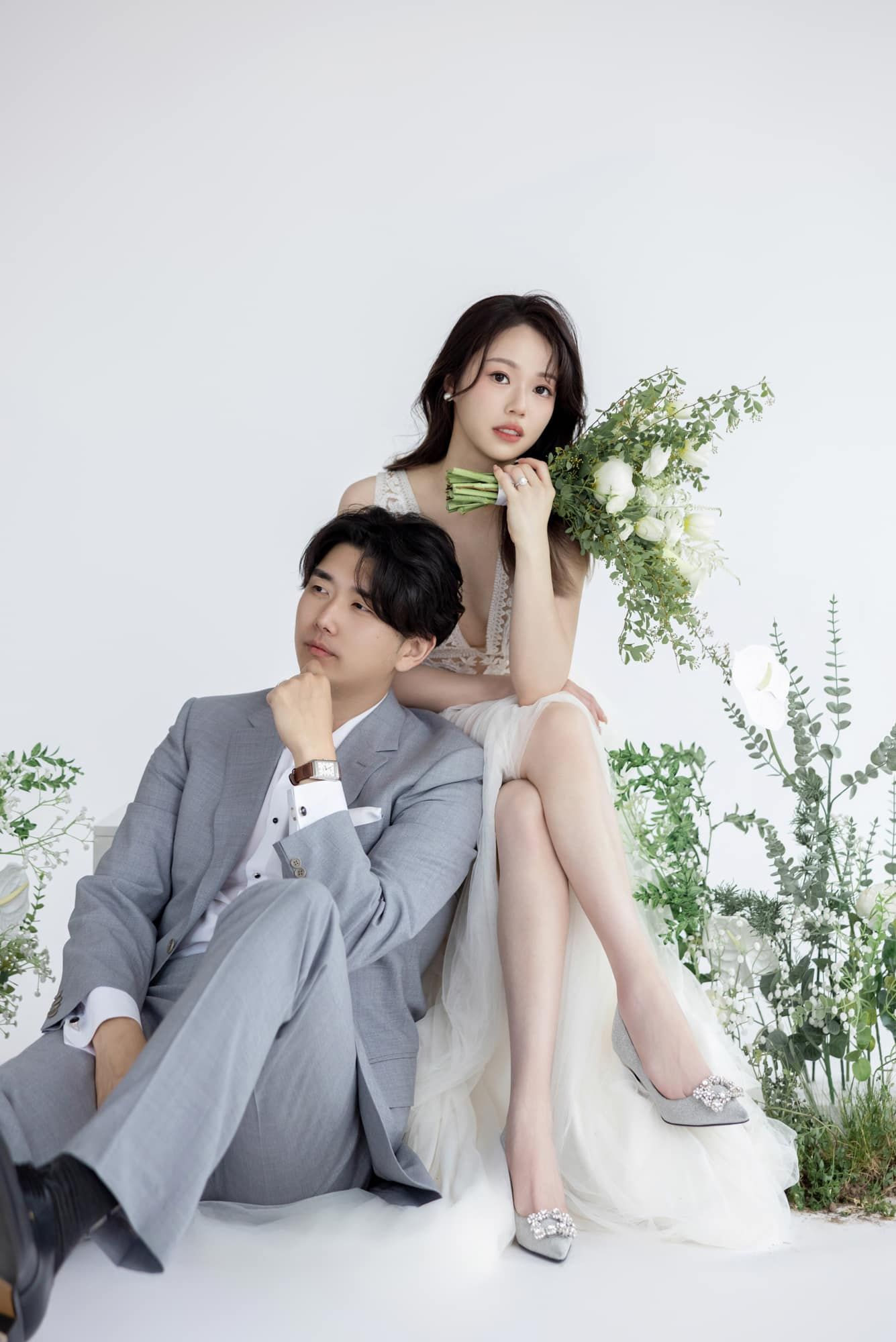 Elegant couple in wedding attire pose with white floral bouquet and greenery in a bright studio setting.
