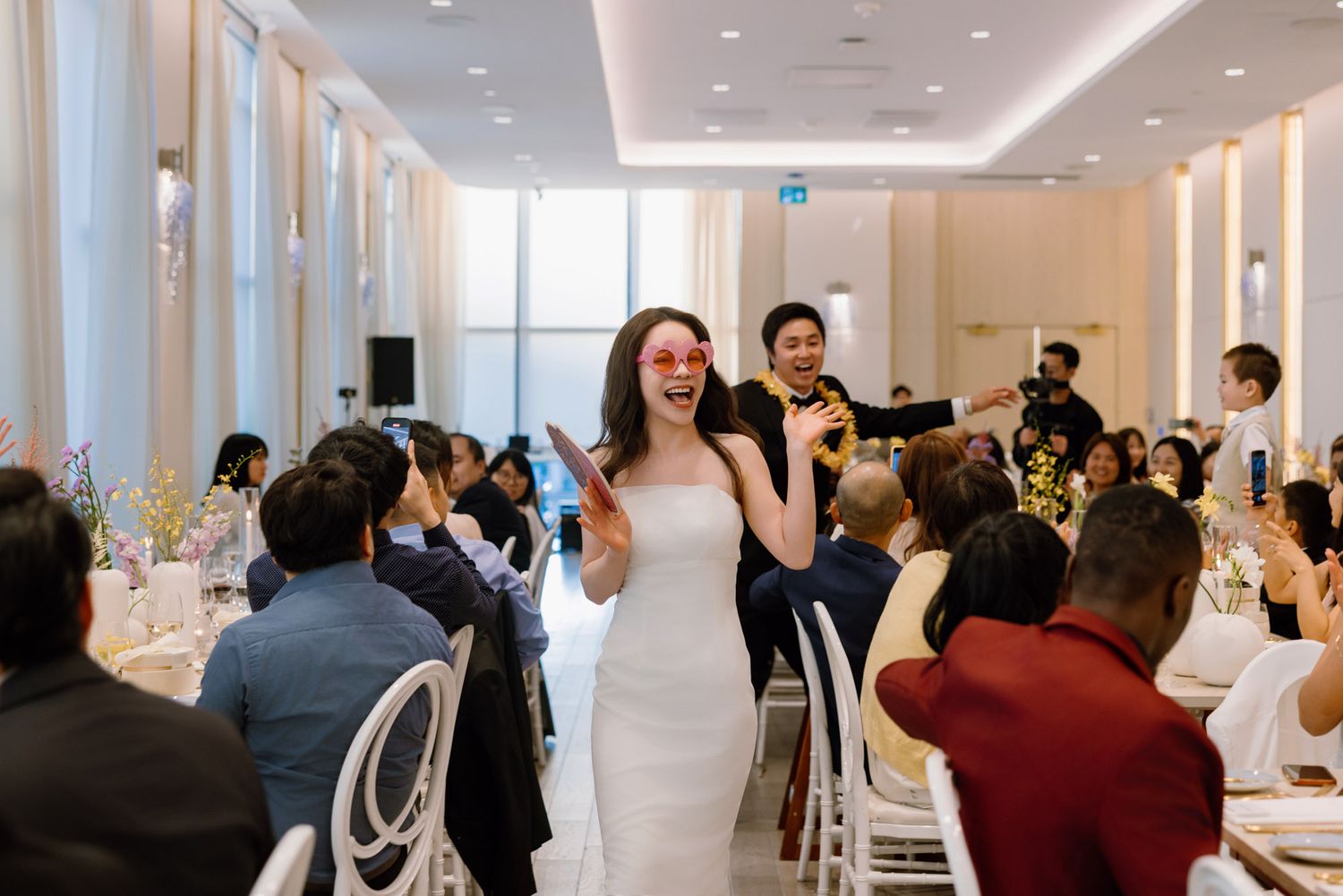 Bride in white gown with fun pink sunglasses celebrating joyfully at wedding reception.