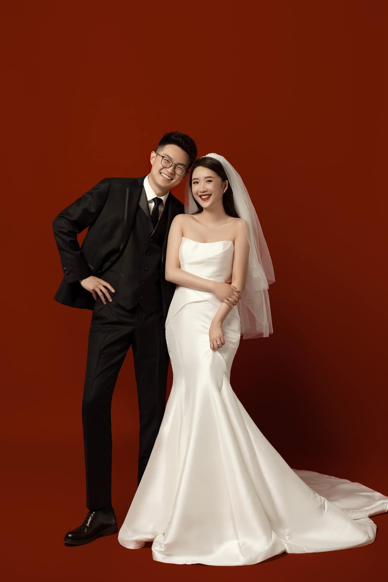 A couple poses for wedding photos, groom in black suit and bride in white mermaid gown with veil on red background.