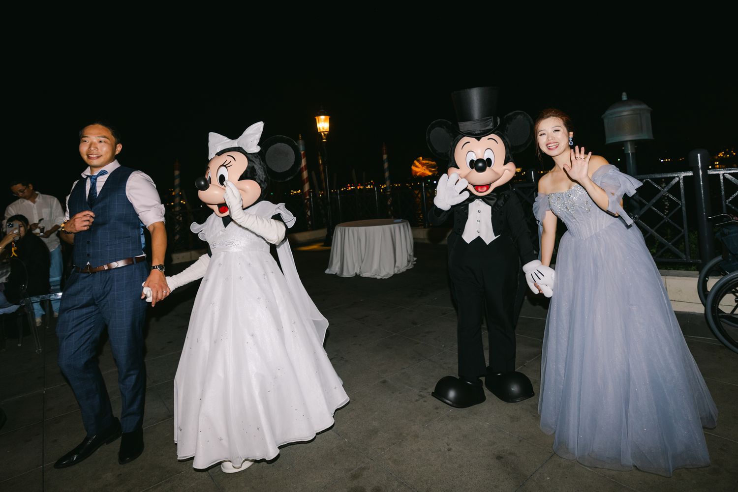 Wedding couple poses with Mickey and Minnie Mouse characters at nighttime Disney celebration.