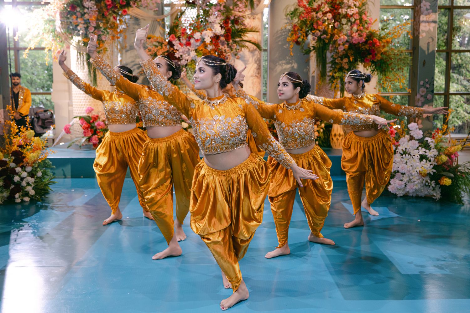 Dancers in golden-orange costumes perform a synchronized traditional dance on a blue floor surrounded by wedding flower arrangements.