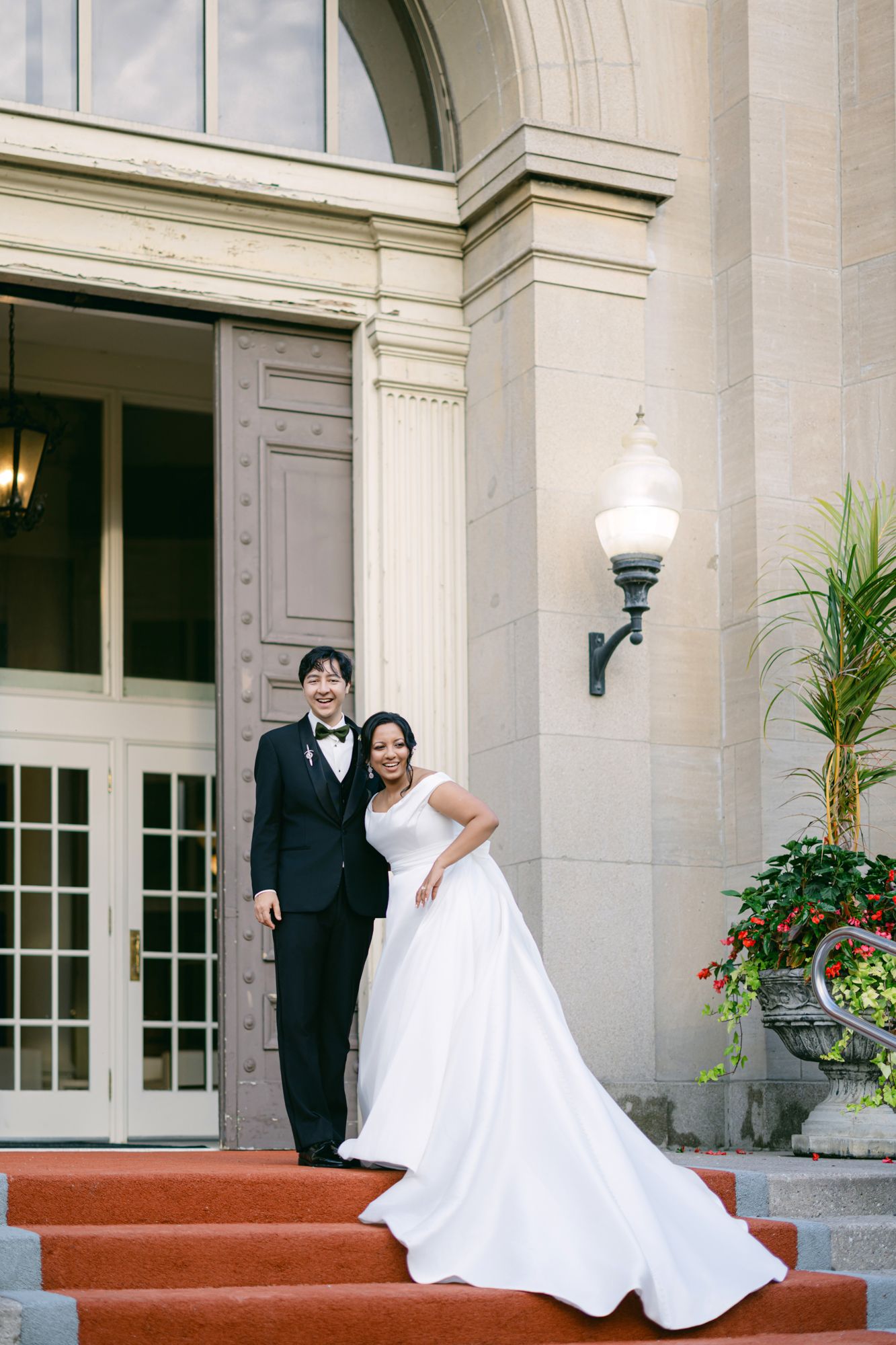 A smiling bride in a flowing white gown and a groom in a black tuxedo pose on red-carpeted steps outside a stately building.