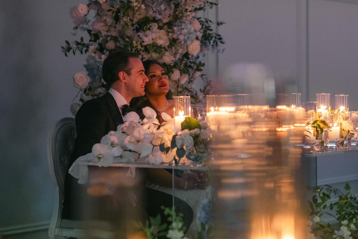Bride and groom sit intimately at candlelit sweetheart table surrounded by white floral arrangements.