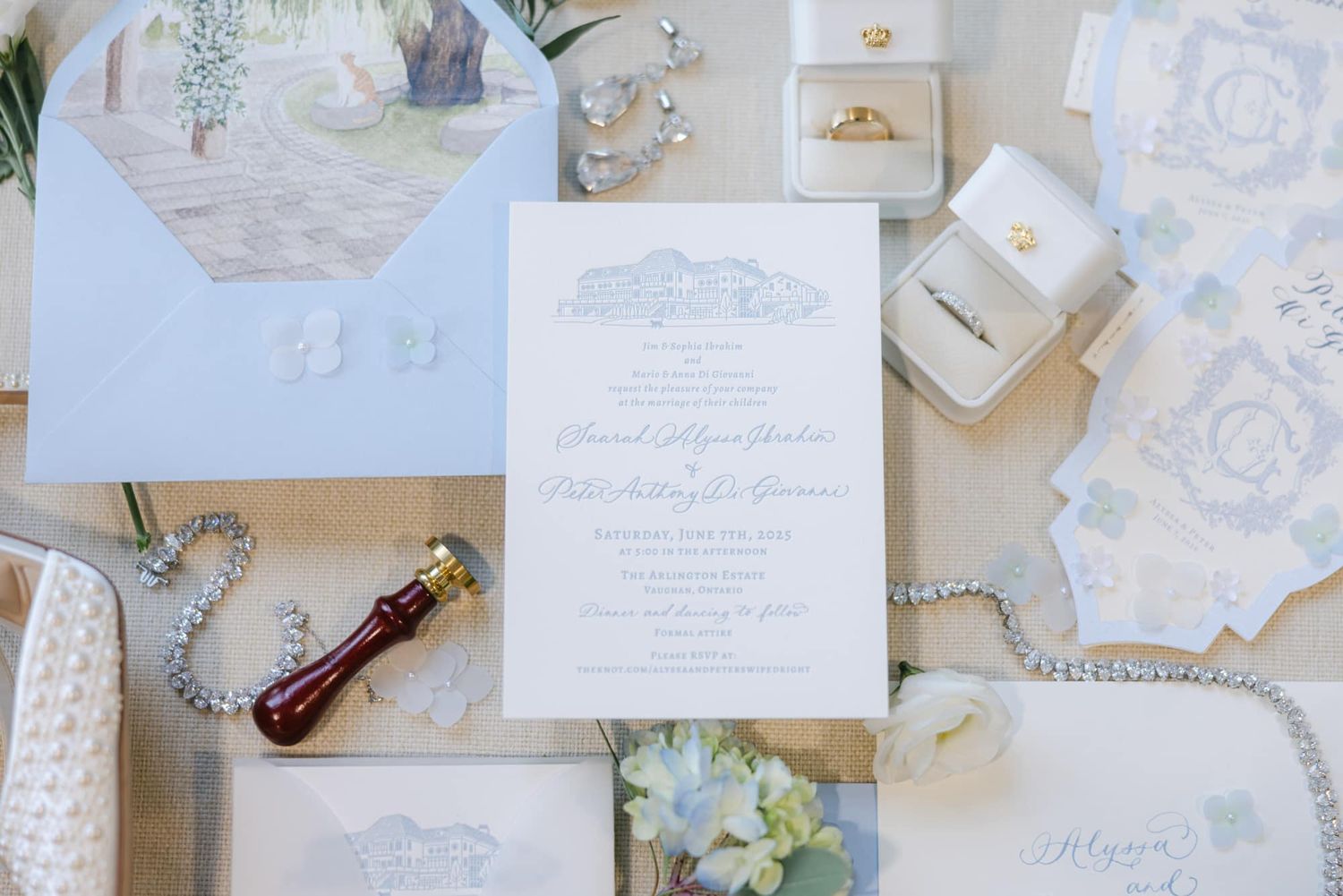 Luxury wedding stationery flatlay featuring blue envelope, gold rings, and delicate white flowers.