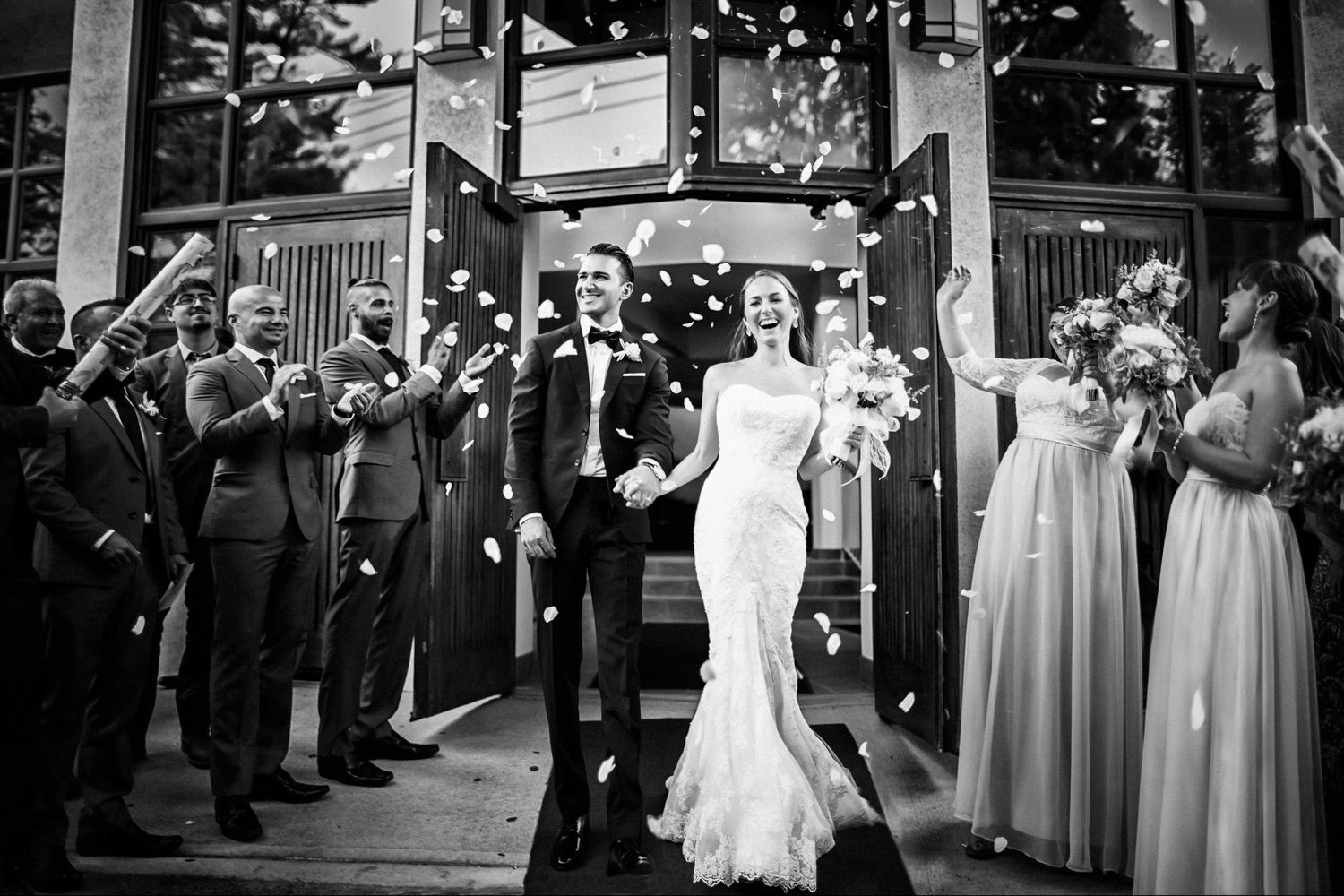Newlyweds exit church doors as guests shower them with white confetti in black and white photo.