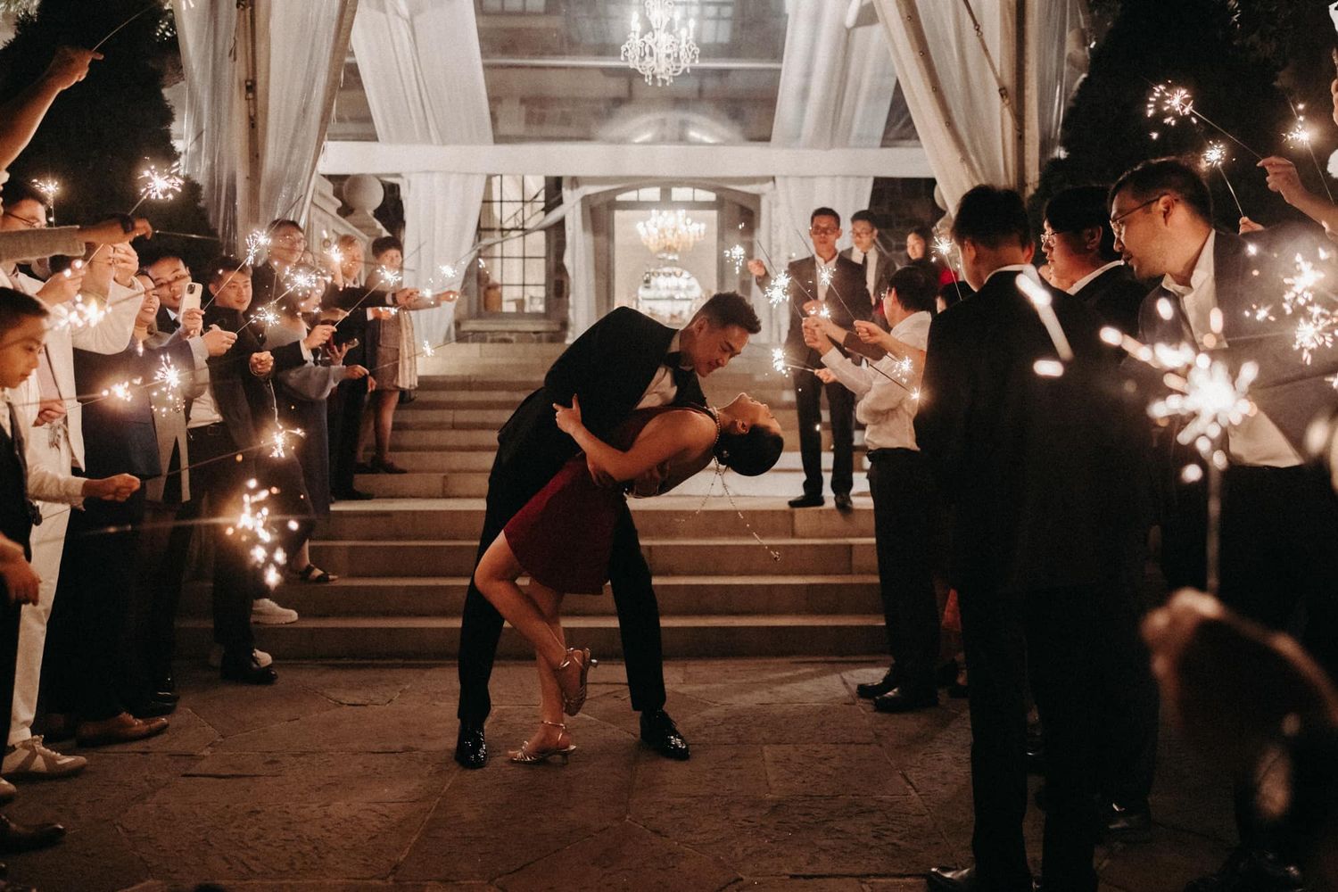 Couple sharing romantic dip kiss while guests line sparkler exit at night wedding celebration.