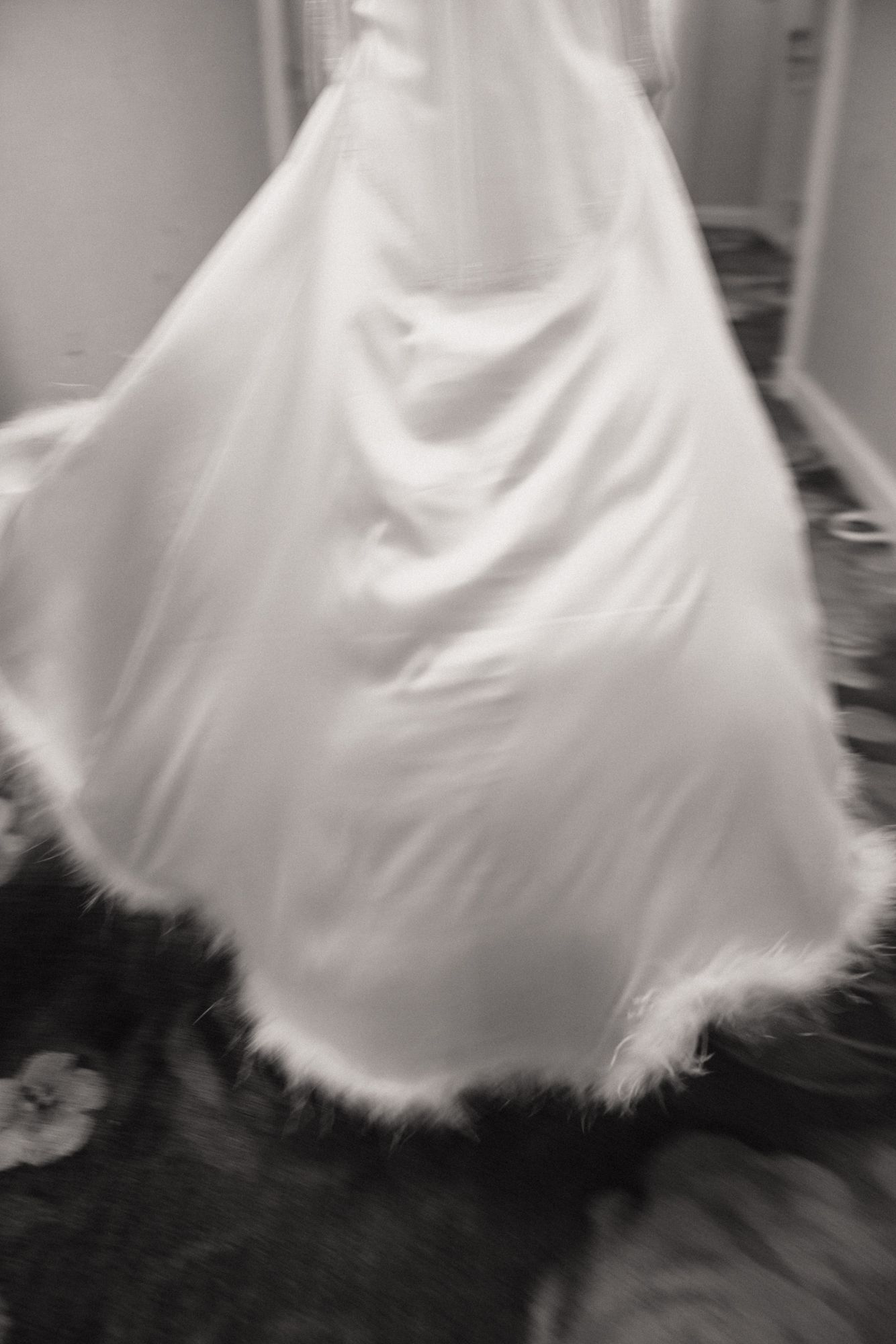 Black and white photo of a flowing white wedding dress with feather trim hem, captured in motion indoors.