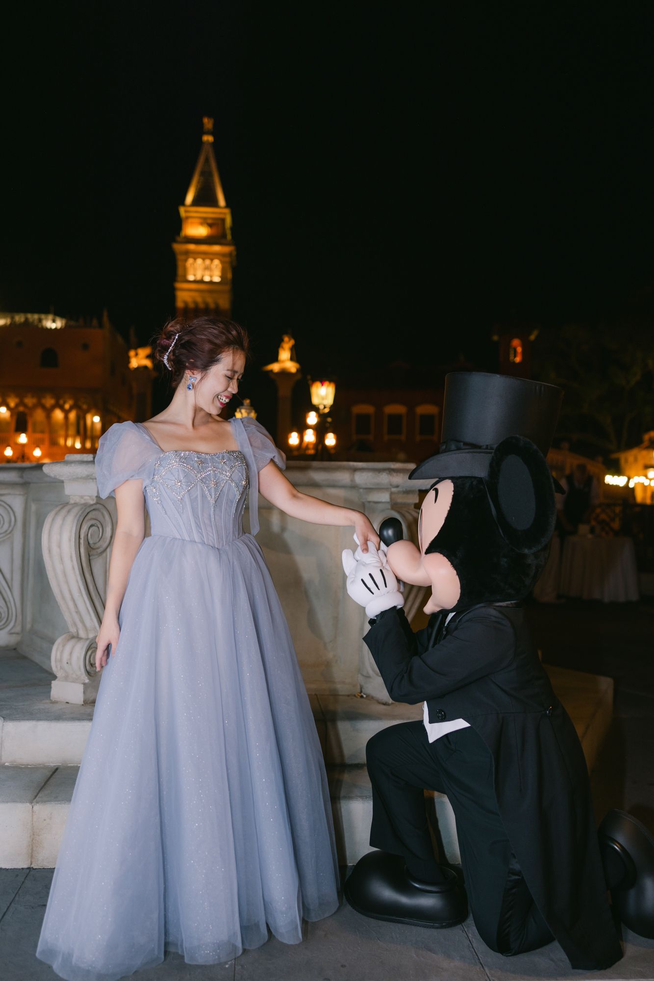 Bride in blue gown poses with tuxedo-clad Mickey Mouse character at Disney World at night.