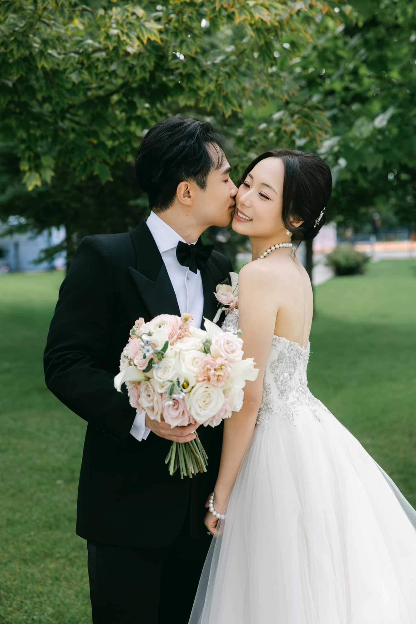 Groom in black tuxedo kisses bride in strapless white gown holding blush bouquet outdoors.