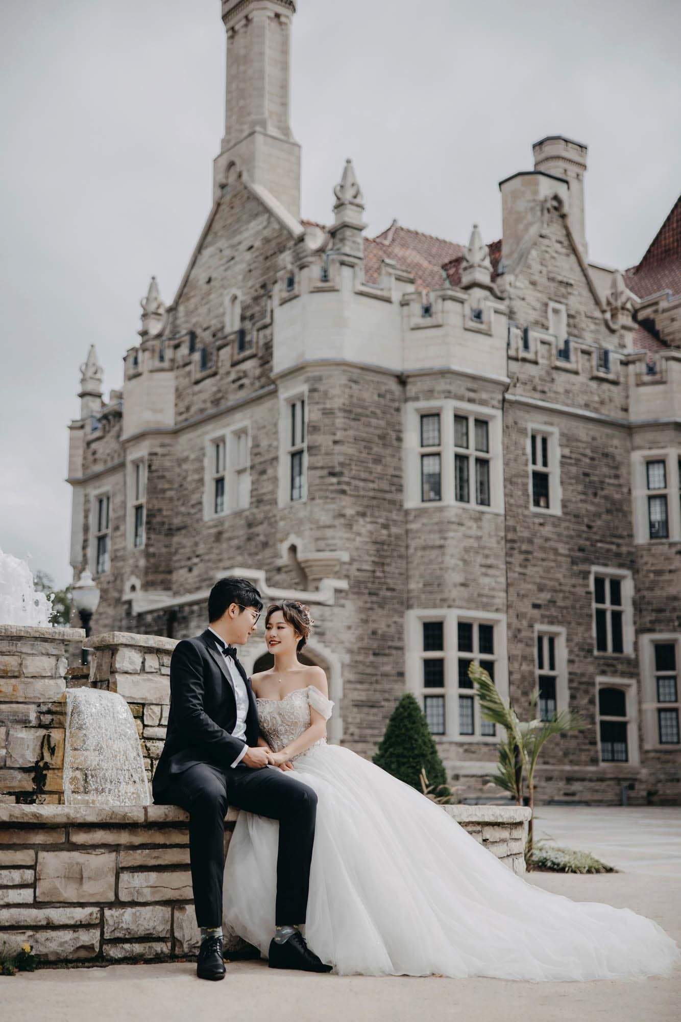 Wedding couple sits together on stone steps before a grand Gothic castle exterior.