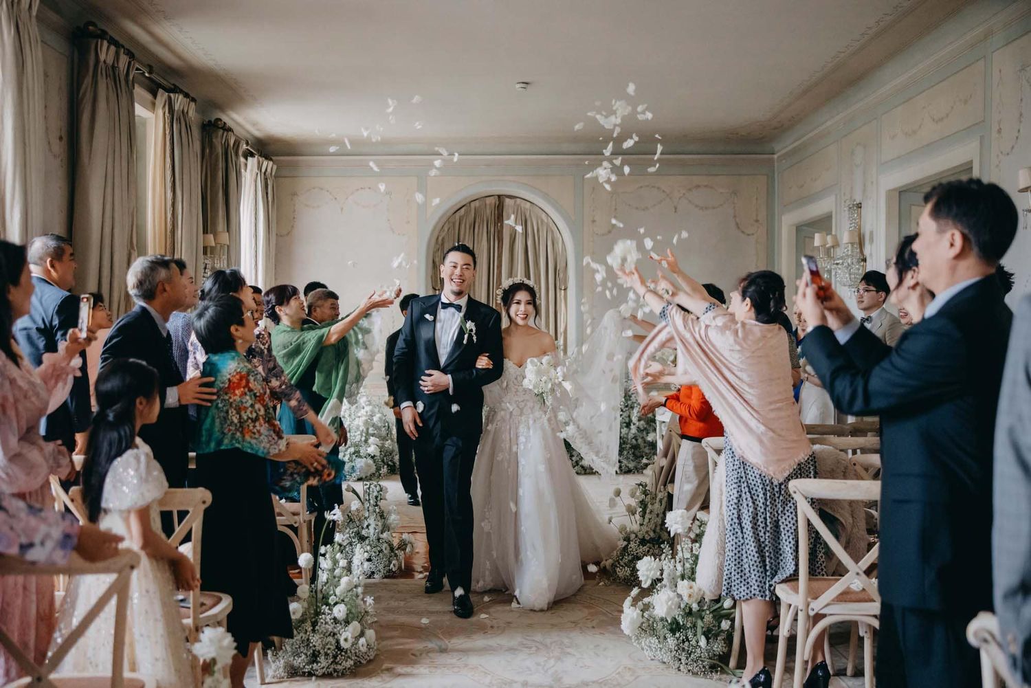 Newlyweds walking down the aisle as guests celebrate with confetti in an elegant floral-decorated venue.