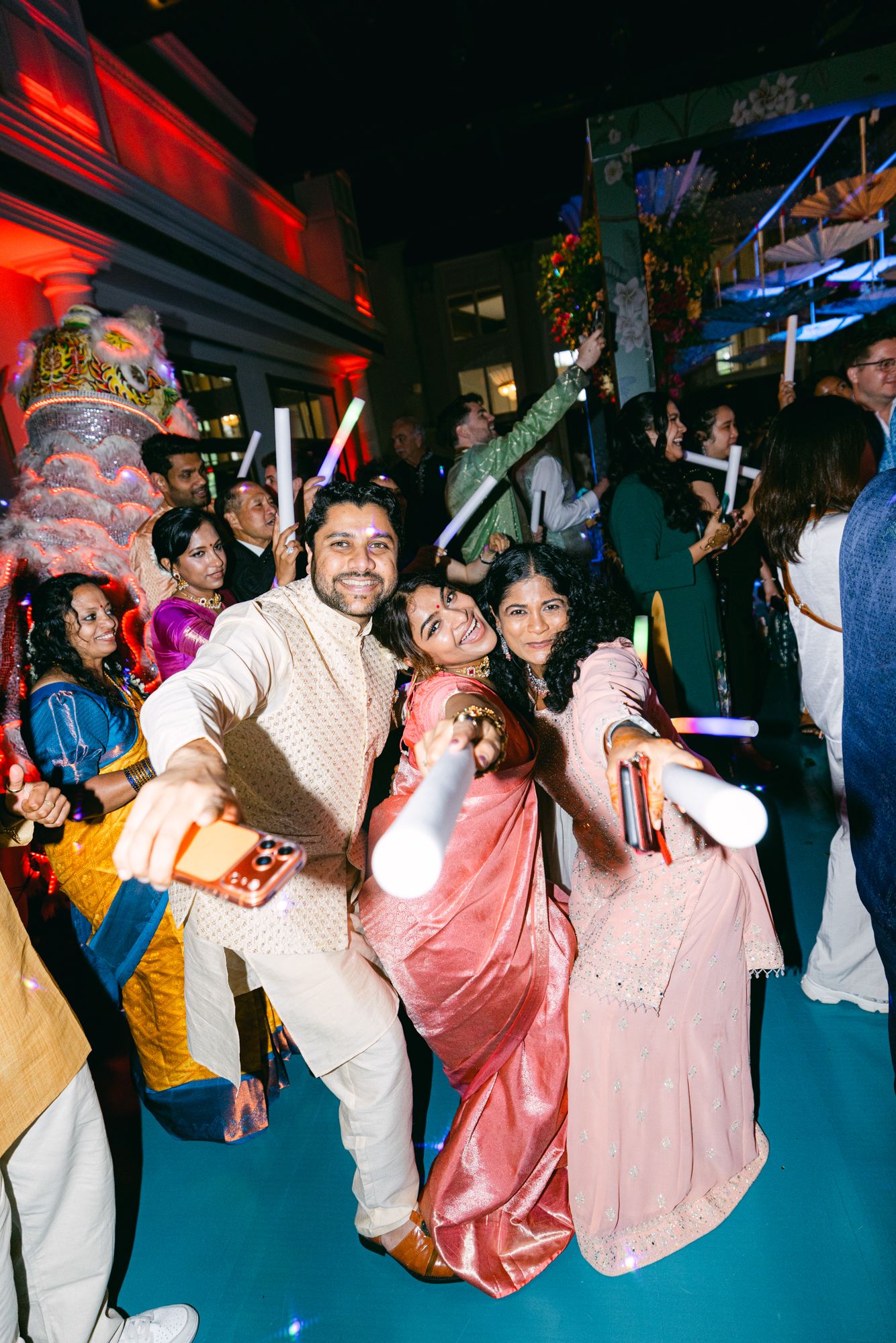 Smiling wedding guests in colorful Indian attire pose with glow sticks while taking a selfie at a lively evening reception.
