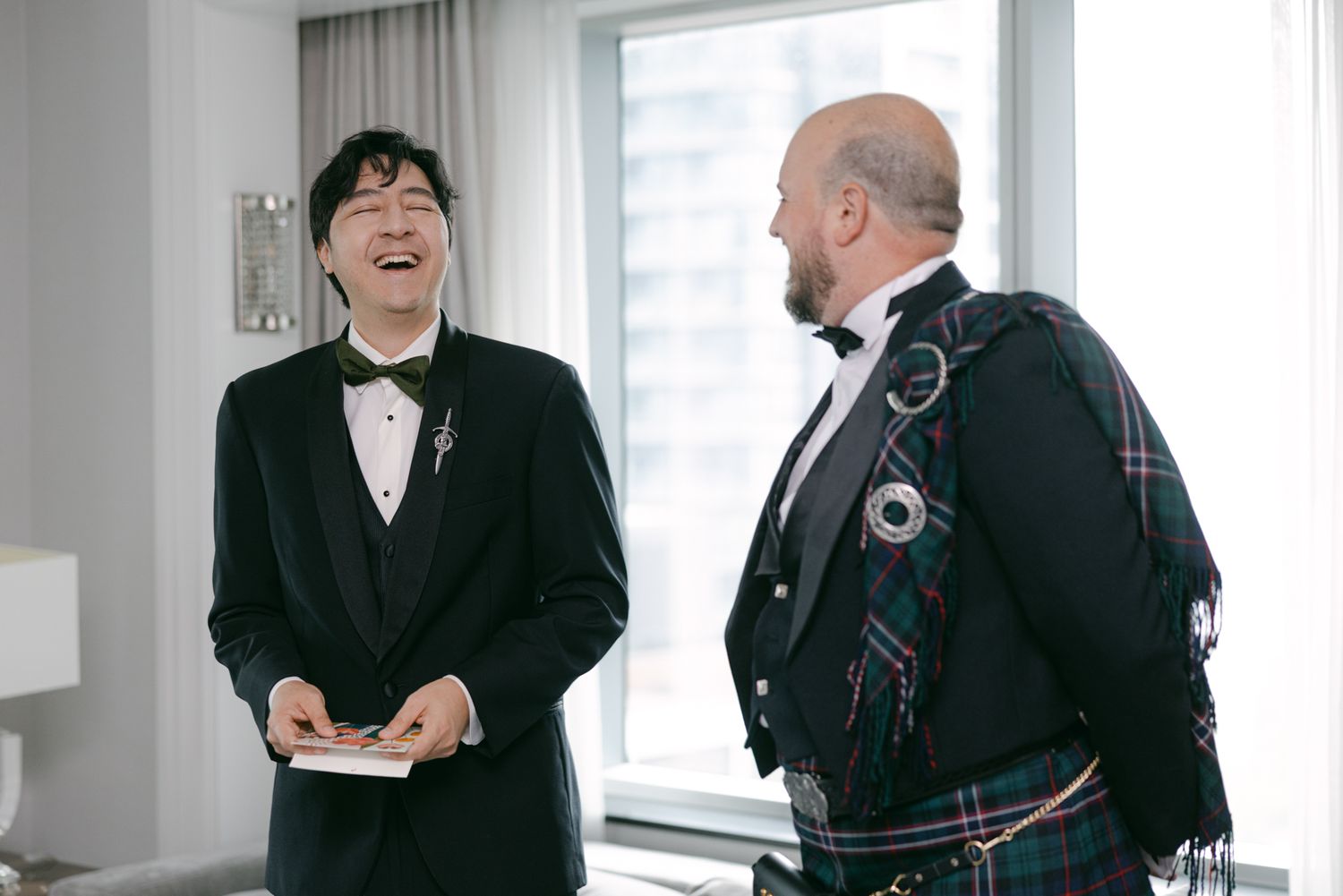 Smiling groom in a black tuxedo and bow tie laughs with a man in a green tartan kilt while getting ready for the wedding.