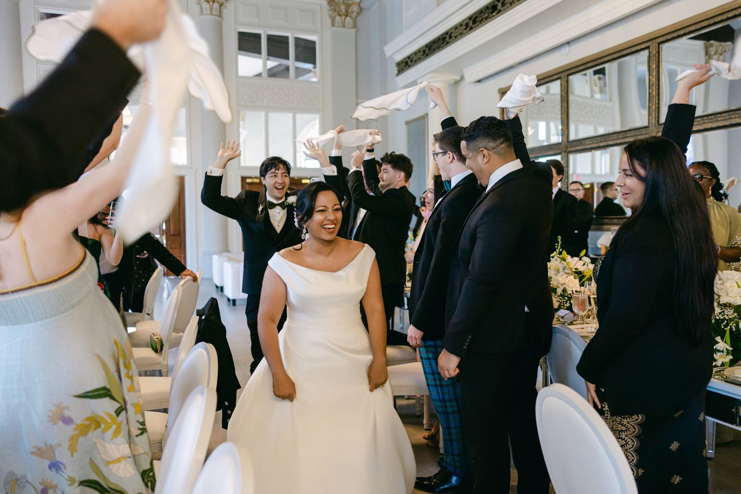 Smiling bride walks toward her table as wedding guests wave white napkins and cheer in celebration at the reception.