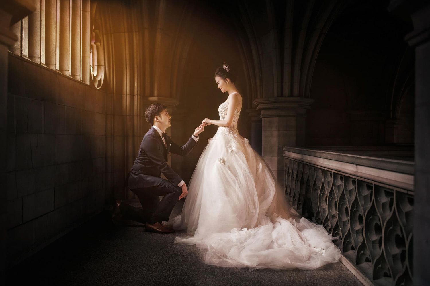 Groom kneeling to place ring on bride's finger in elegant wedding photo with gothic architecture backdrop.