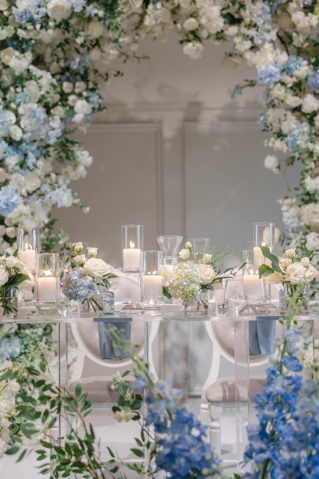 Sweetheart table framed by a lush blue hydrangea and white floral arch with candles and mirror table.