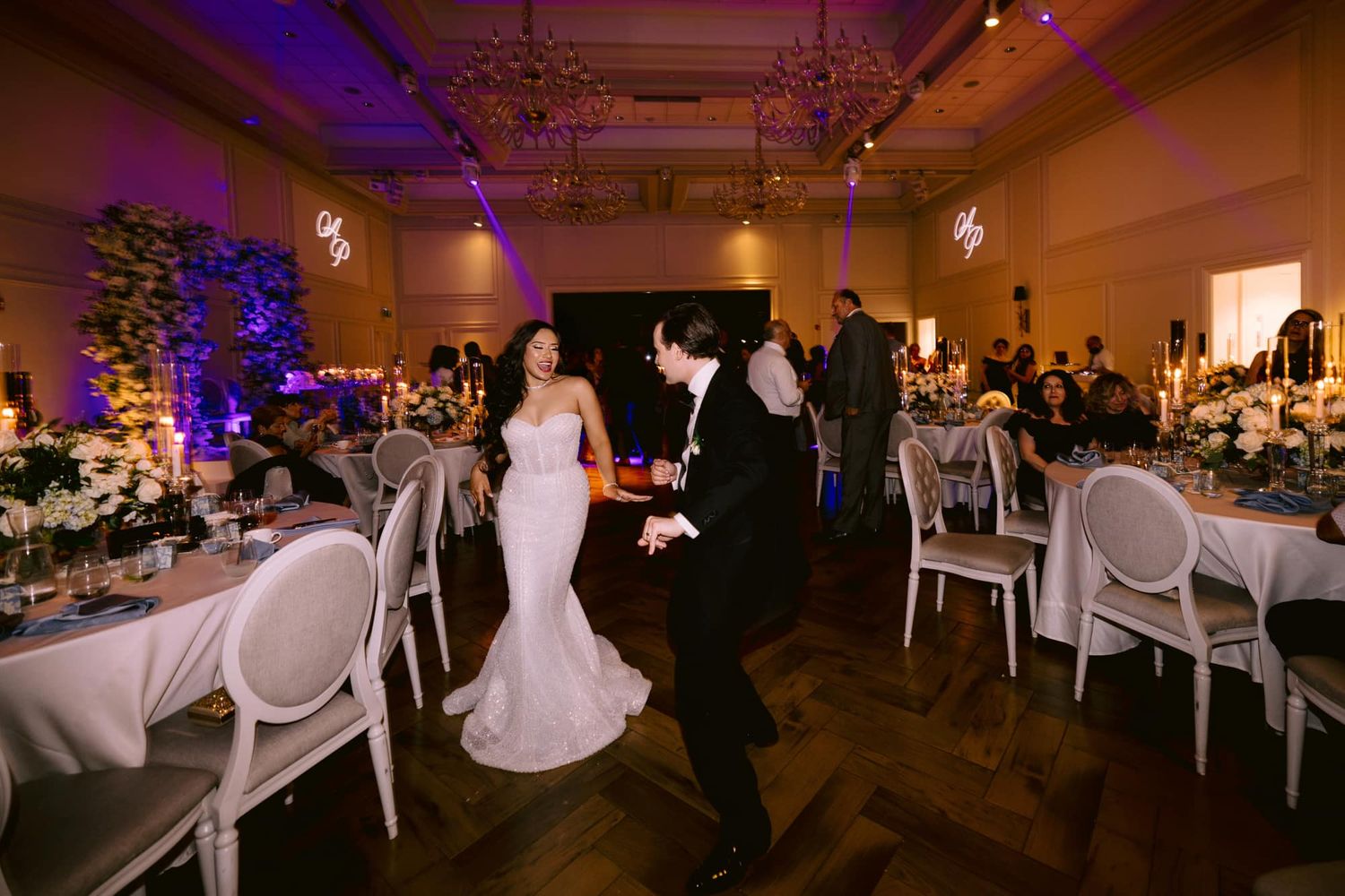 Bride and groom sharing first dance in elegant ballroom with floral arrangements and purple lighting.