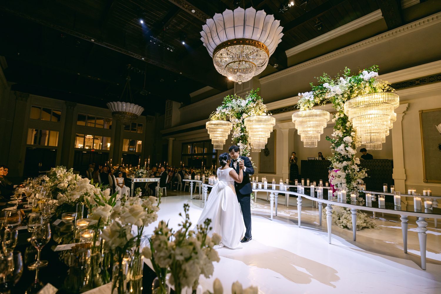 Bride and groom share a first dance on a glossy ballroom floor beneath glowing chandeliers and lush floral arrangements.