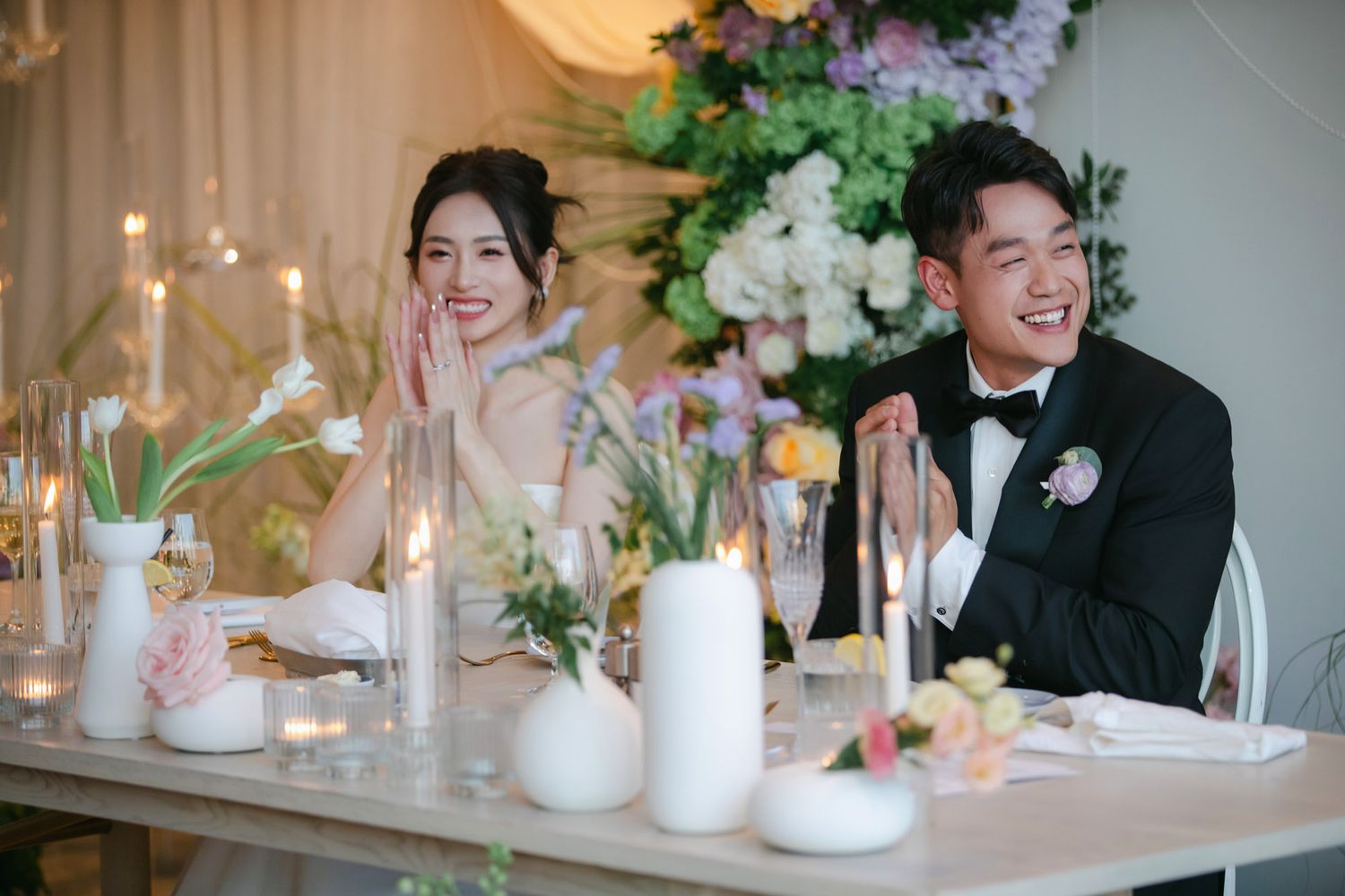 Smiling bride and groom seated at elegant wedding reception table with candles and floral arrangements.