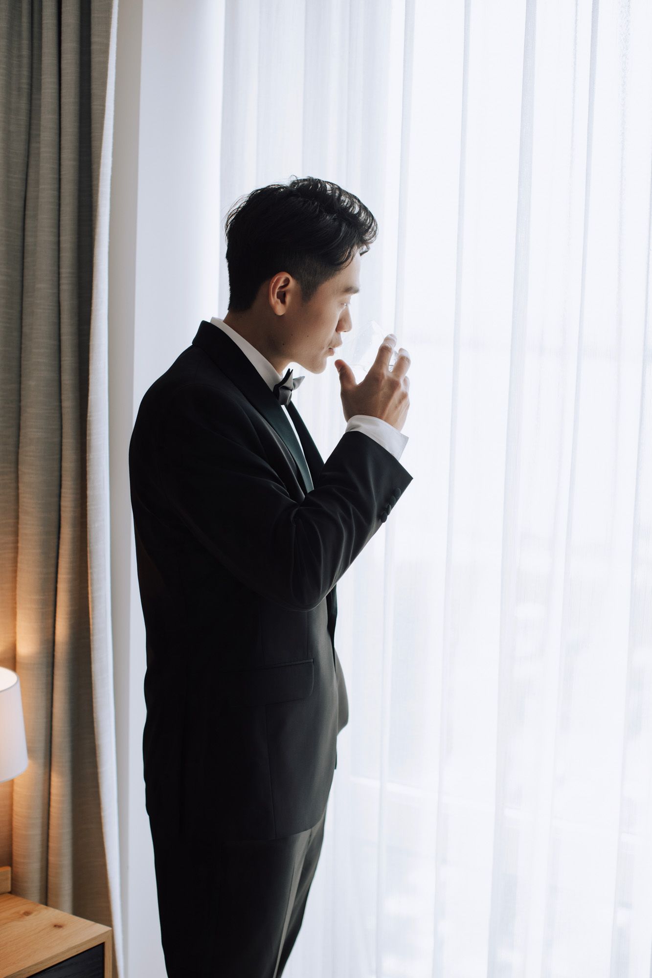 Elegant groom in black tuxedo standing thoughtfully by sheer curtained hotel room window.