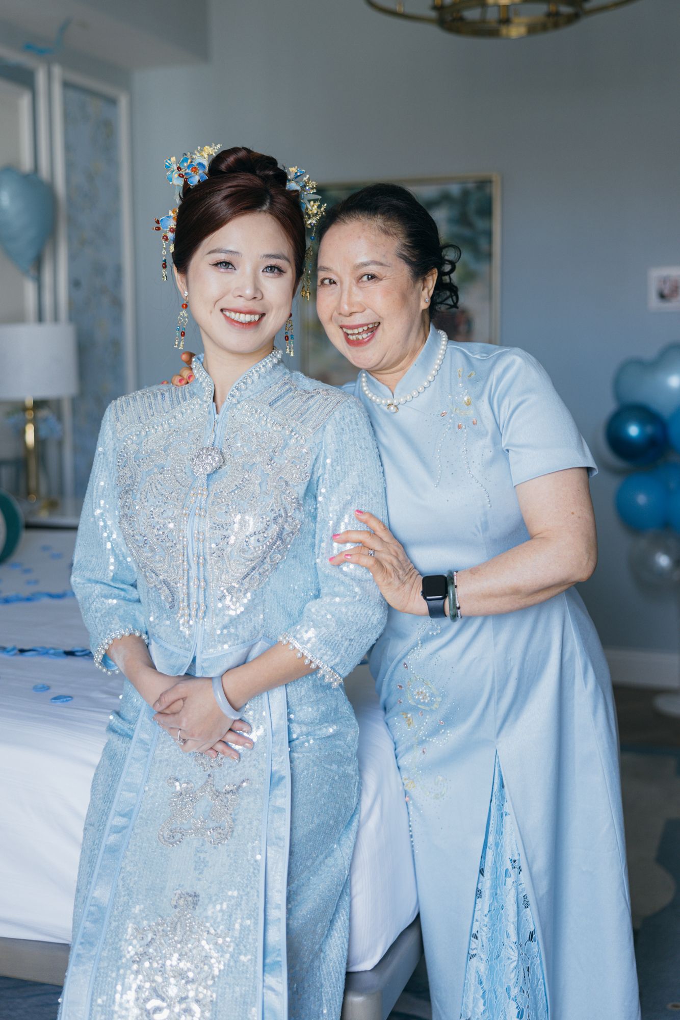 Two women in matching light blue ao dai dresses with floral hair accessories pose together at a wedding celebration.