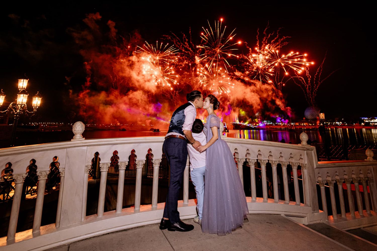 Couple kissing on a balcony with vibrant fireworks lighting the night sky behind them.
